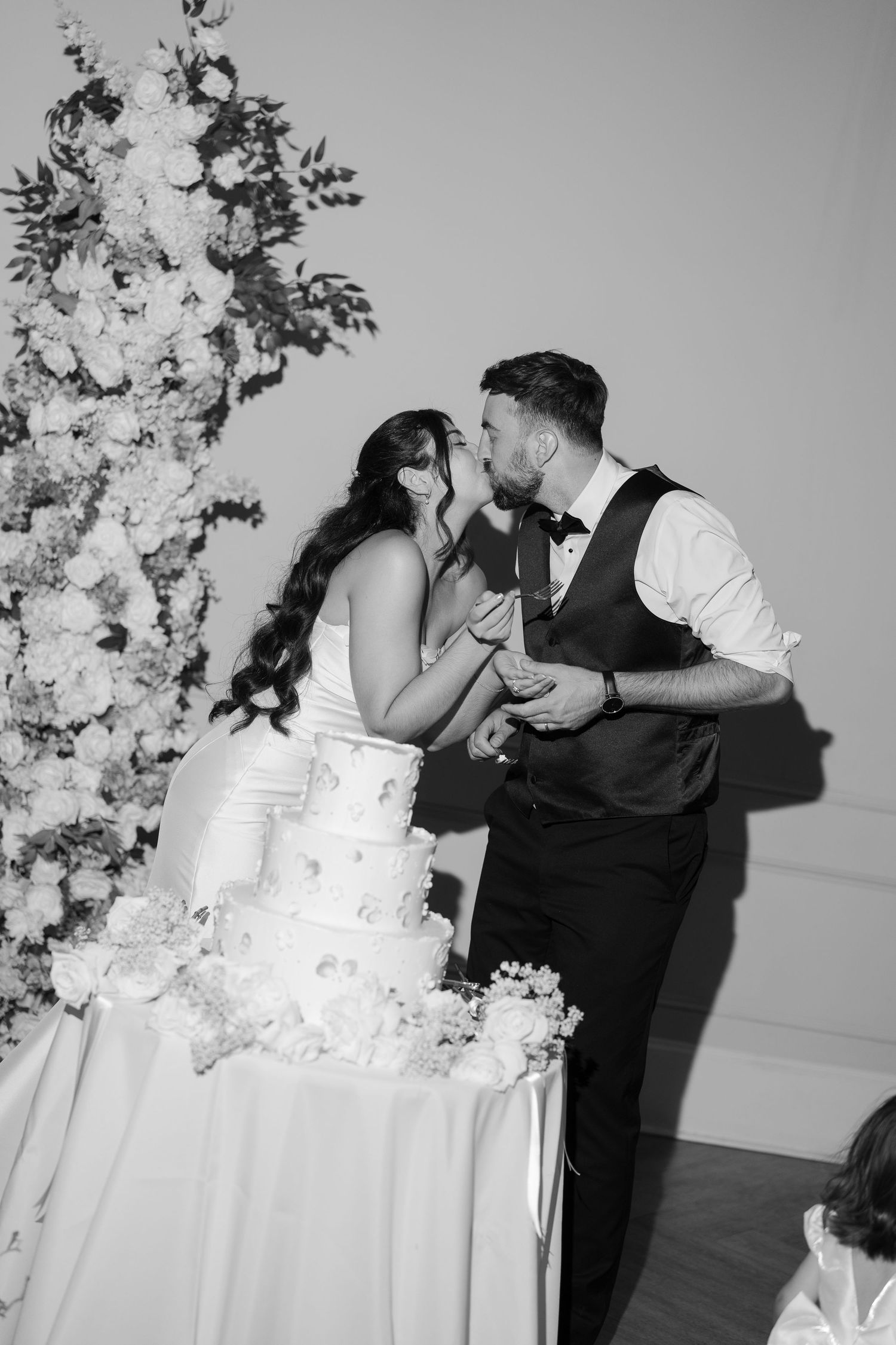 Black and white photo of bride and groom kissing while cutting tiered wedding cake.