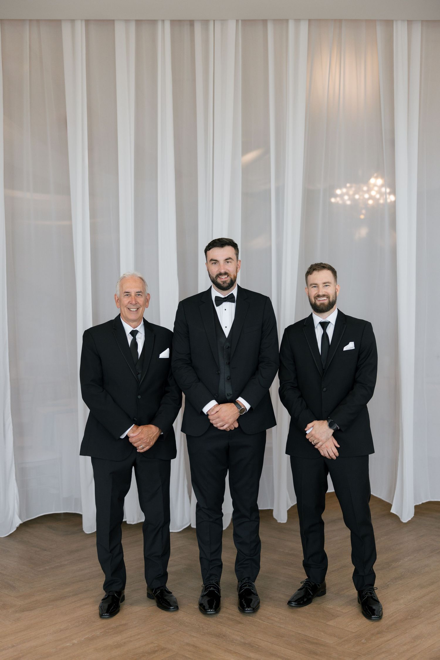 Three groomsmen in black tuxedos pose together in front of white draping at a wedding venue.