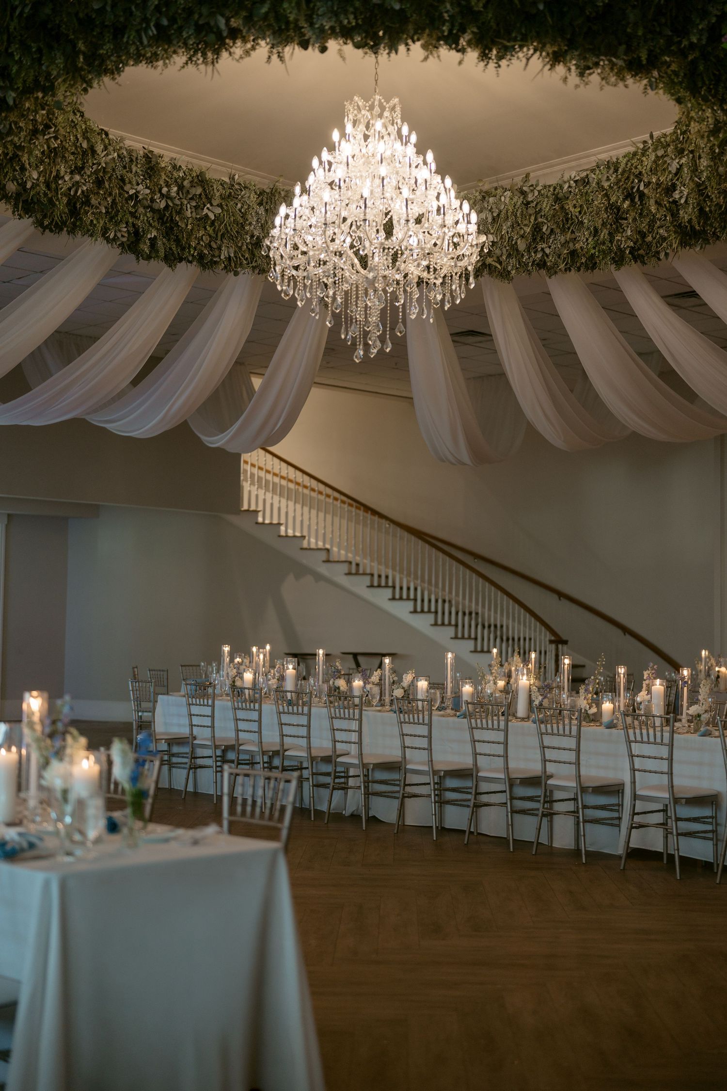 Elegant wedding reception hall with crystal chandelier, draped ceilings, greenery, and long candlelit tables.