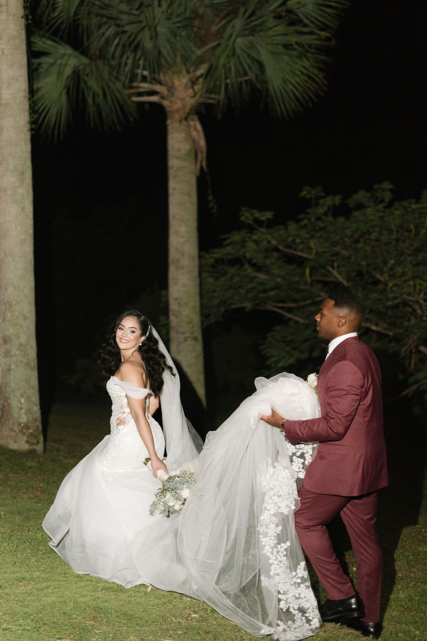 A couple in wedding attire poses on a lawn at night, with the groom holding the bride's dress train.