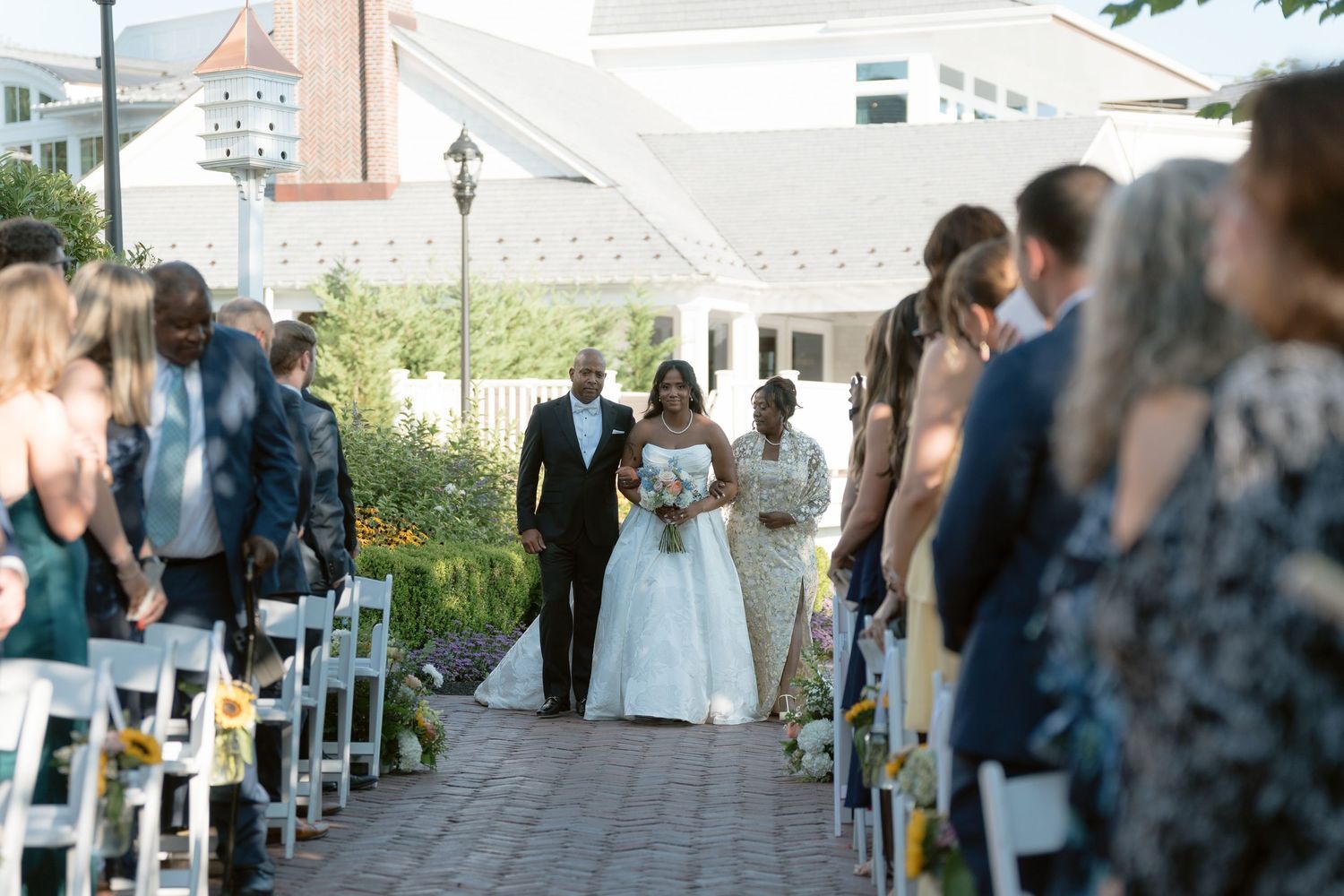 Bride walks down the aisle with family at an outdoor wedding ceremony with white chairs and floral decorations.