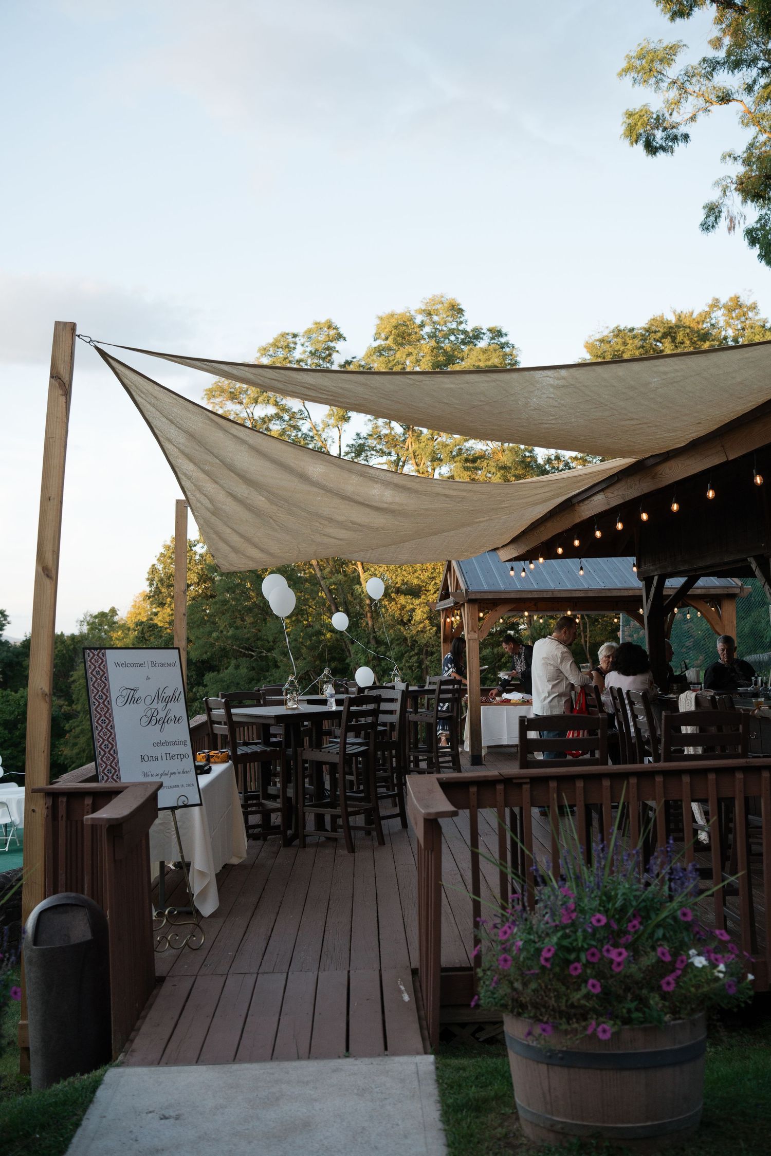 Outdoor wedding venue deck with sail shade canopy, string lights, bistro tables, and a barrel planter with purple flowers.