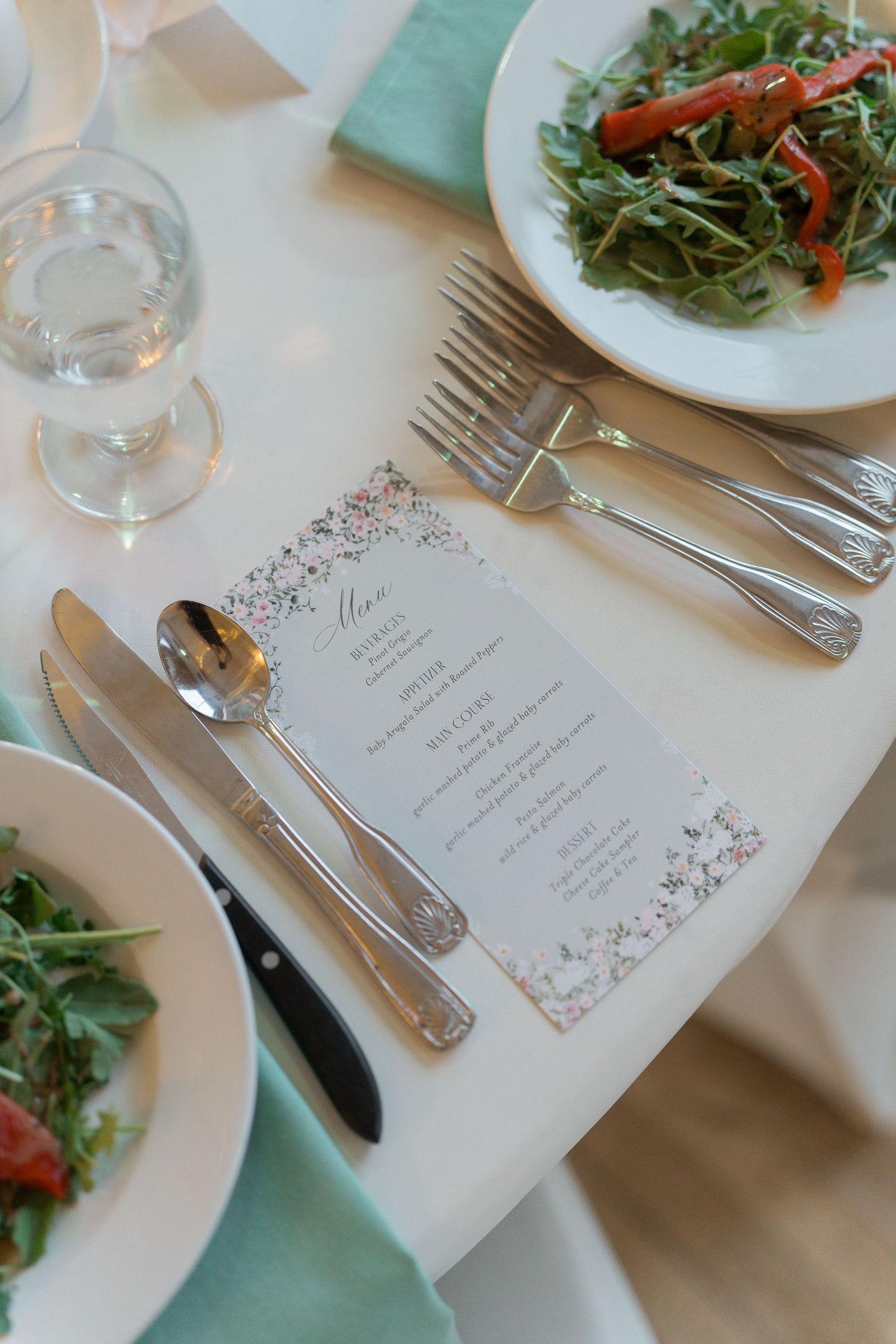 Elegant wedding reception place setting with silver cutlery, floral menu card, and fresh arugula salad plates.