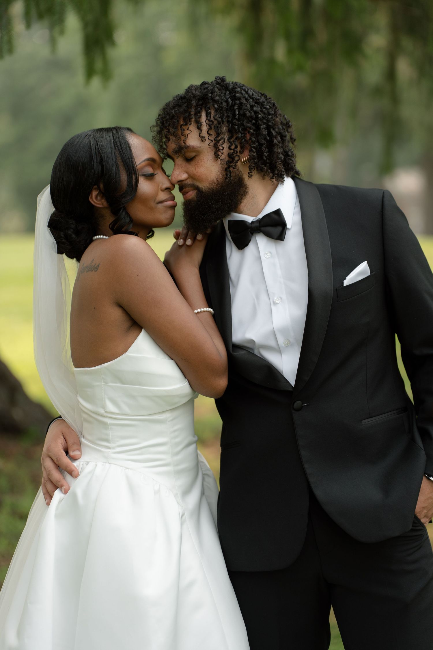 Black couple sharing romantic moment outdoors, bride in white satin gown and groom in black tuxedo with bow tie.