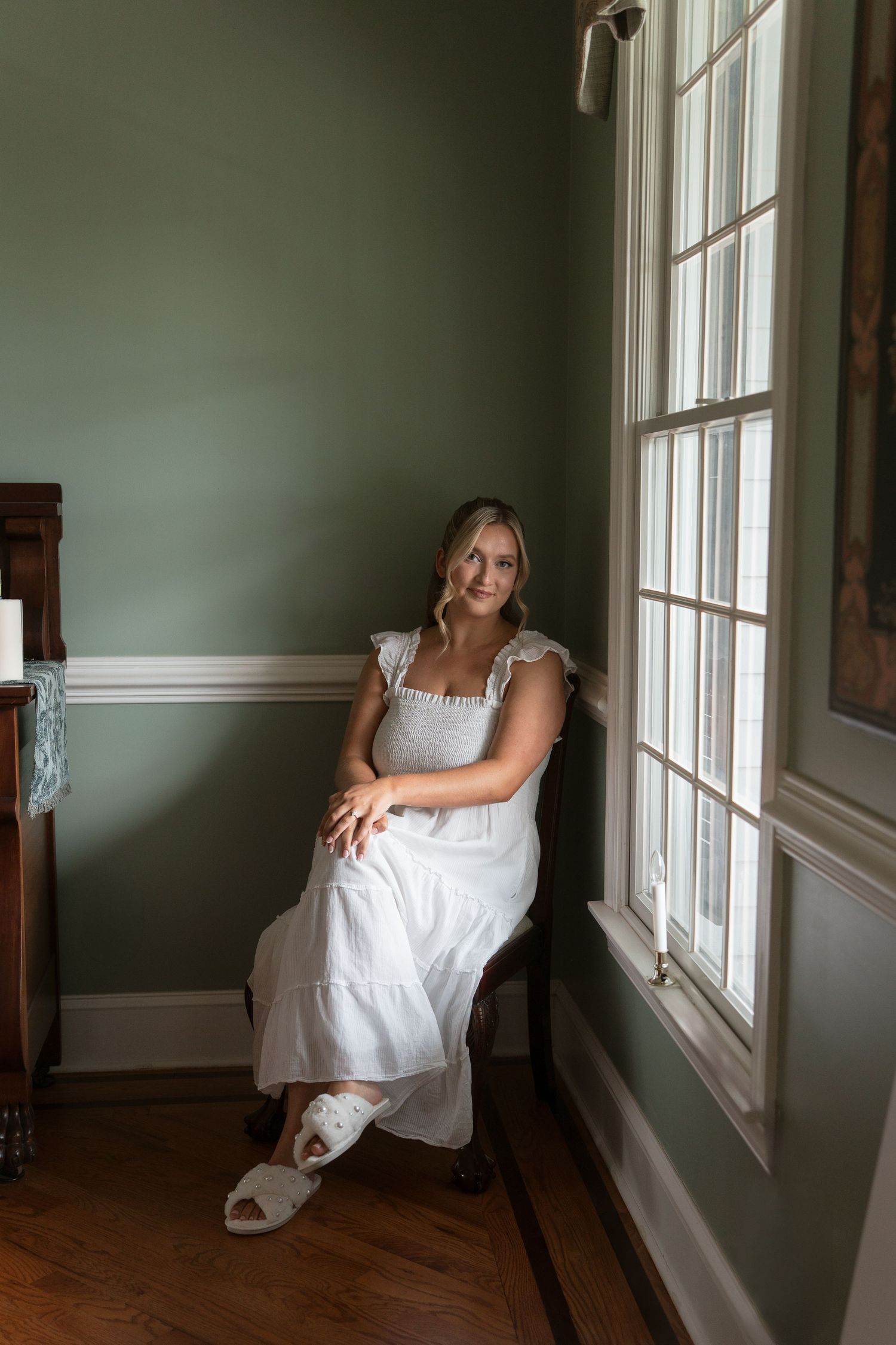 A young woman in a white dress and sandals sits gracefully in a chair by a window in a green room.