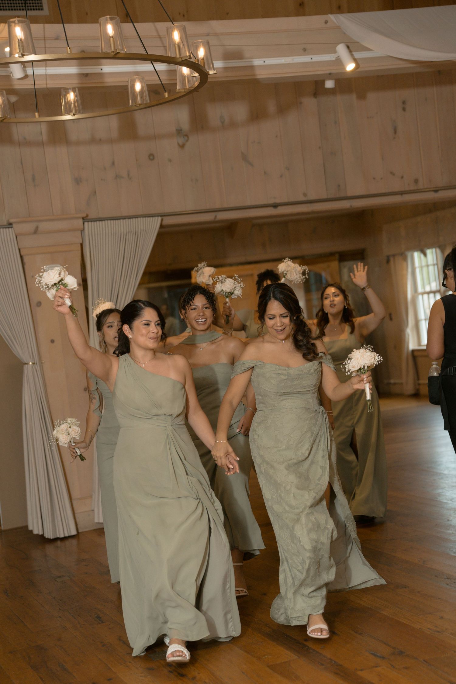 Bridesmaids in sage green gowns walk joyfully holding white bouquets in a rustic wood-paneled wedding venue.