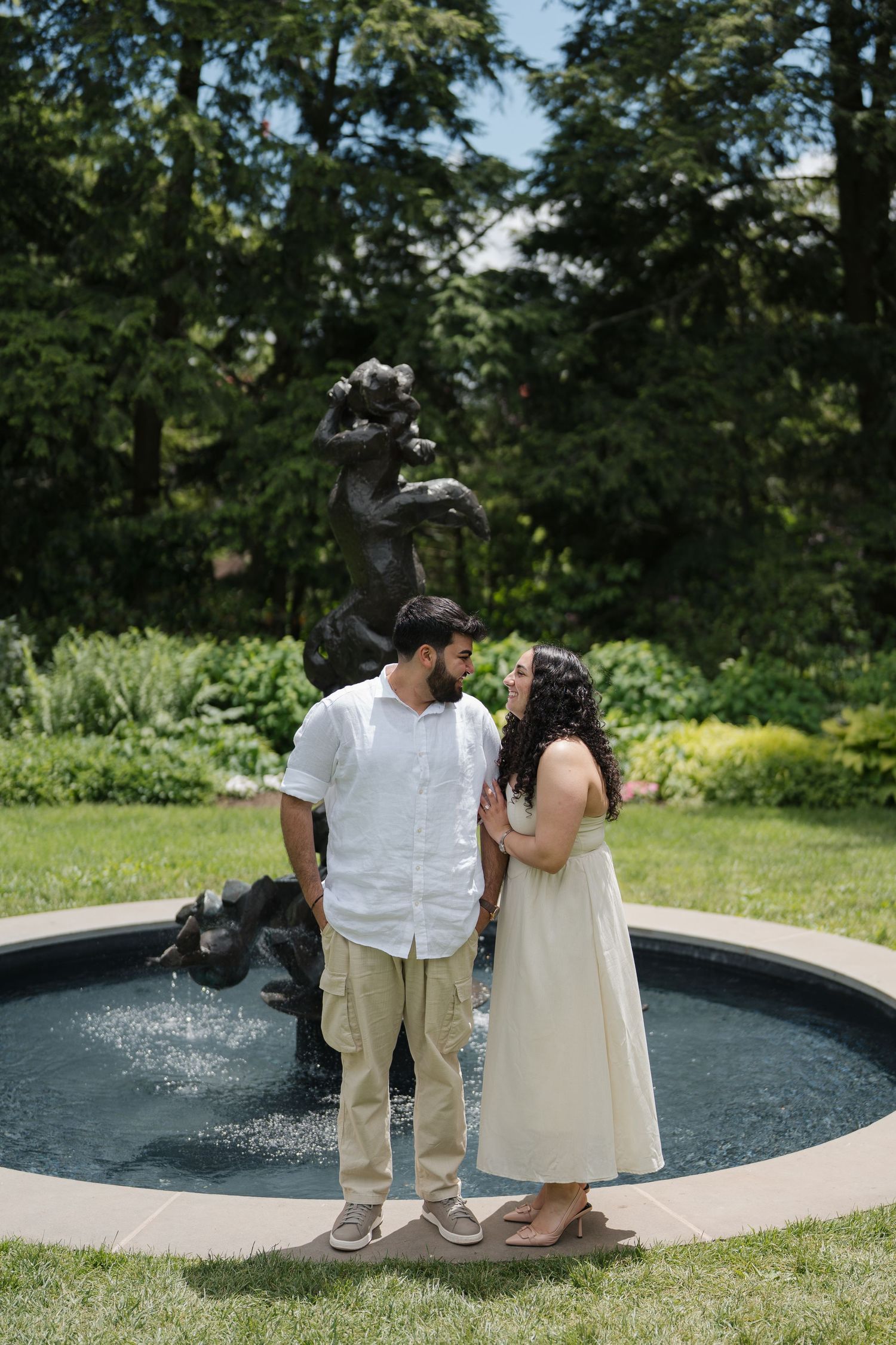 Couple standing by a garden fountain with a dark sculpture, surrounded by lush greenery on a sunny day.