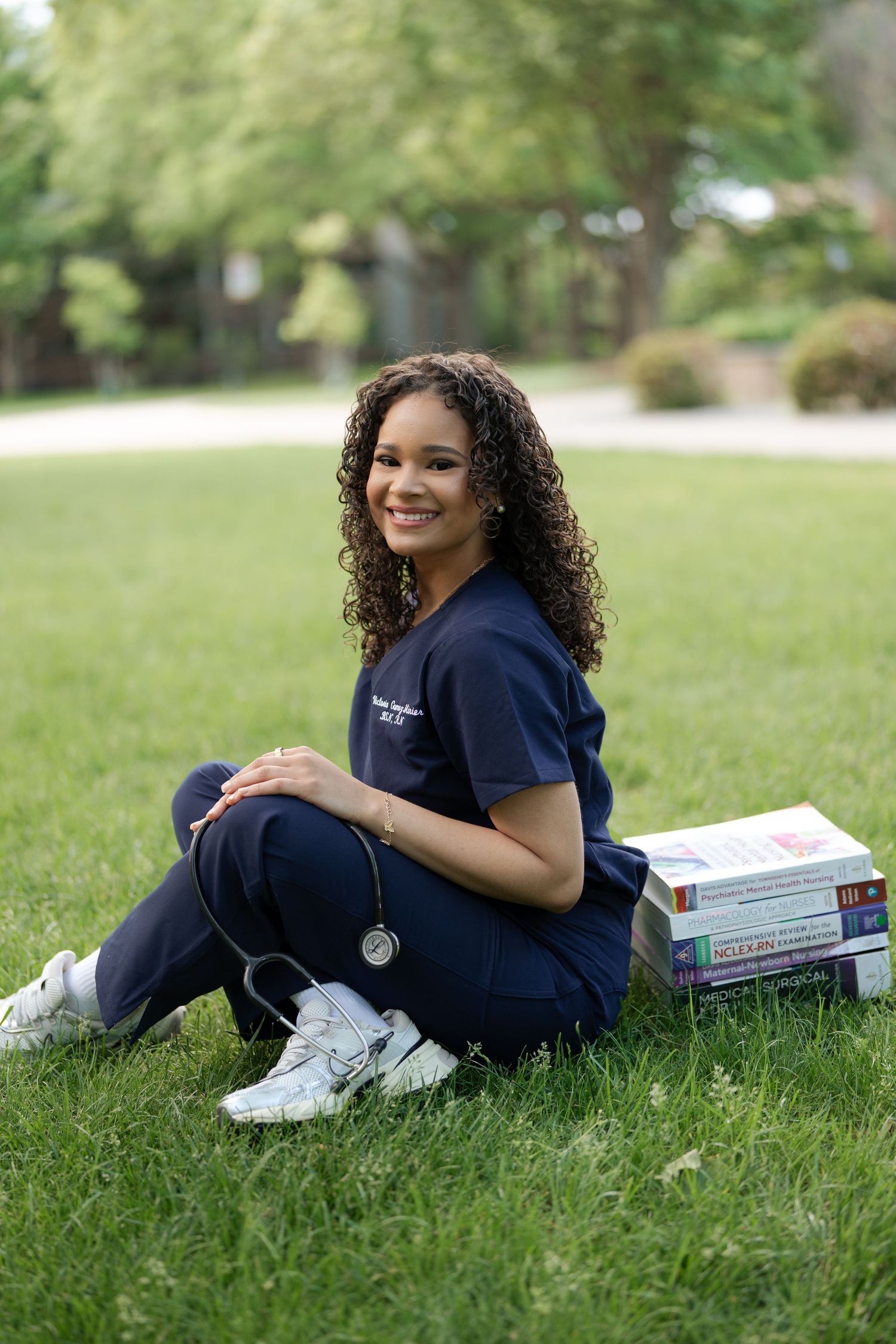 Female nursing student in navy scrubs sitting on grass with stacked medical textbooks and stethoscope.