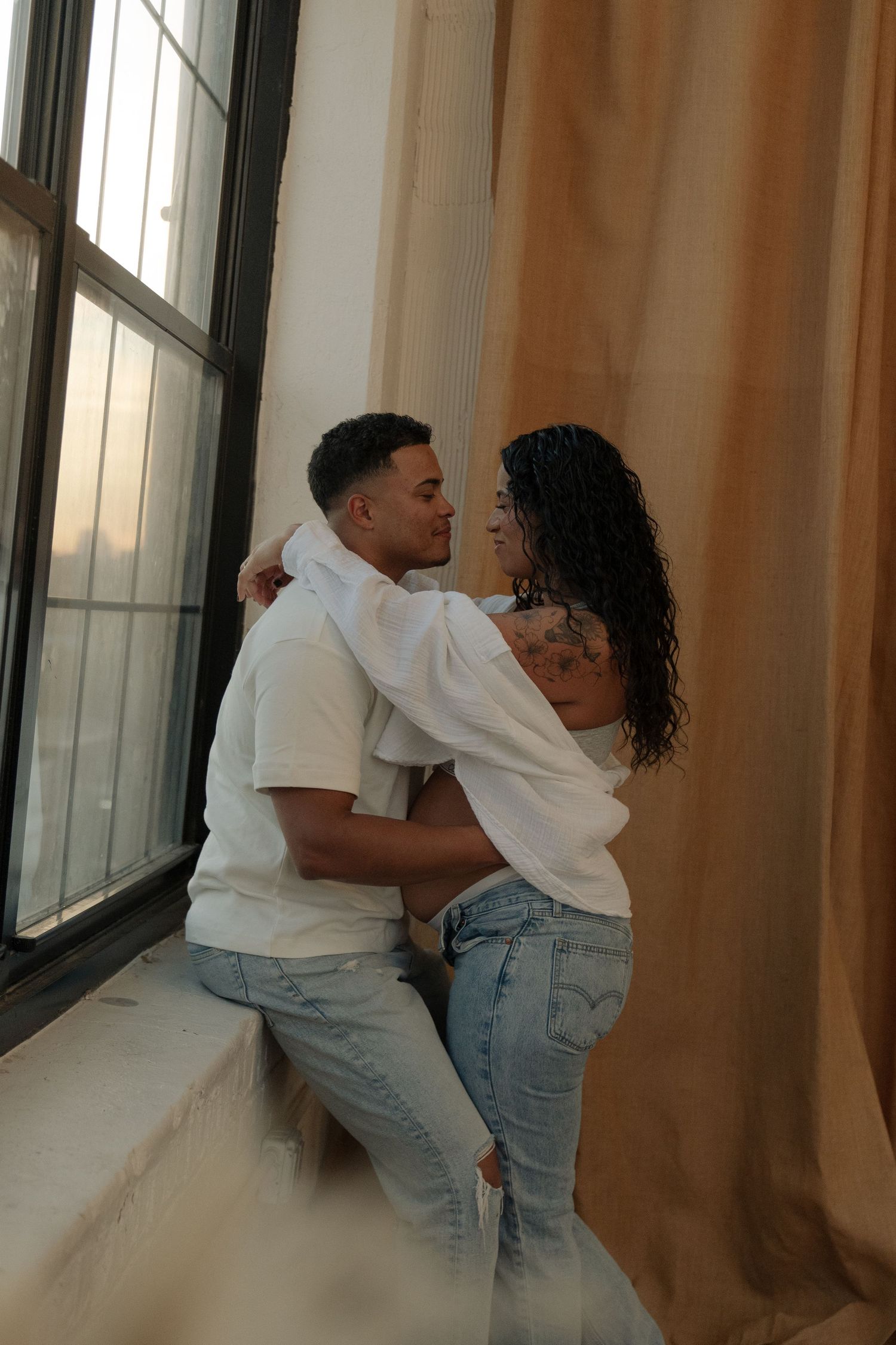 Couple embracing romantically by a large window with warm curtains, wearing matching white shirts and jeans.