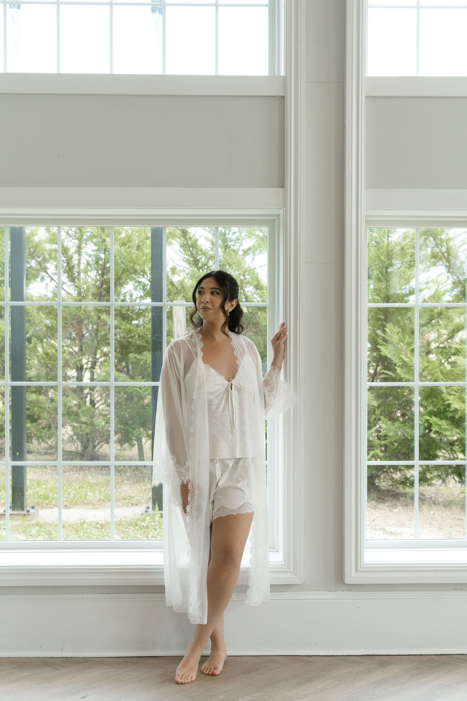 Bride in white lace robe and bridal lingerie standing by large windows with garden views.