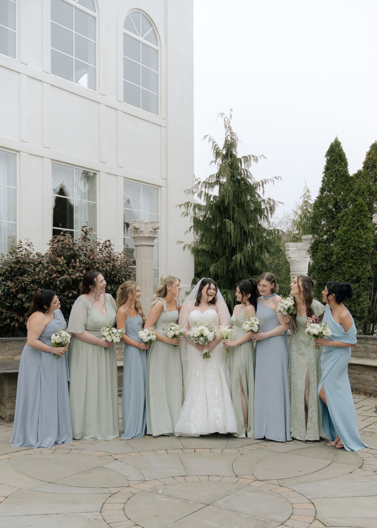 Wedding party in light blue and cream dresses pose in a curved line formation on stone patio during outdoor celebration.