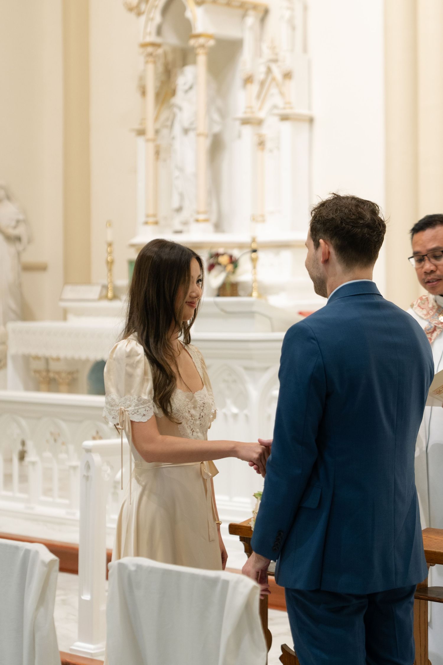 A couple holds hands during their intimate church wedding ceremony with ornate white altar in the background.