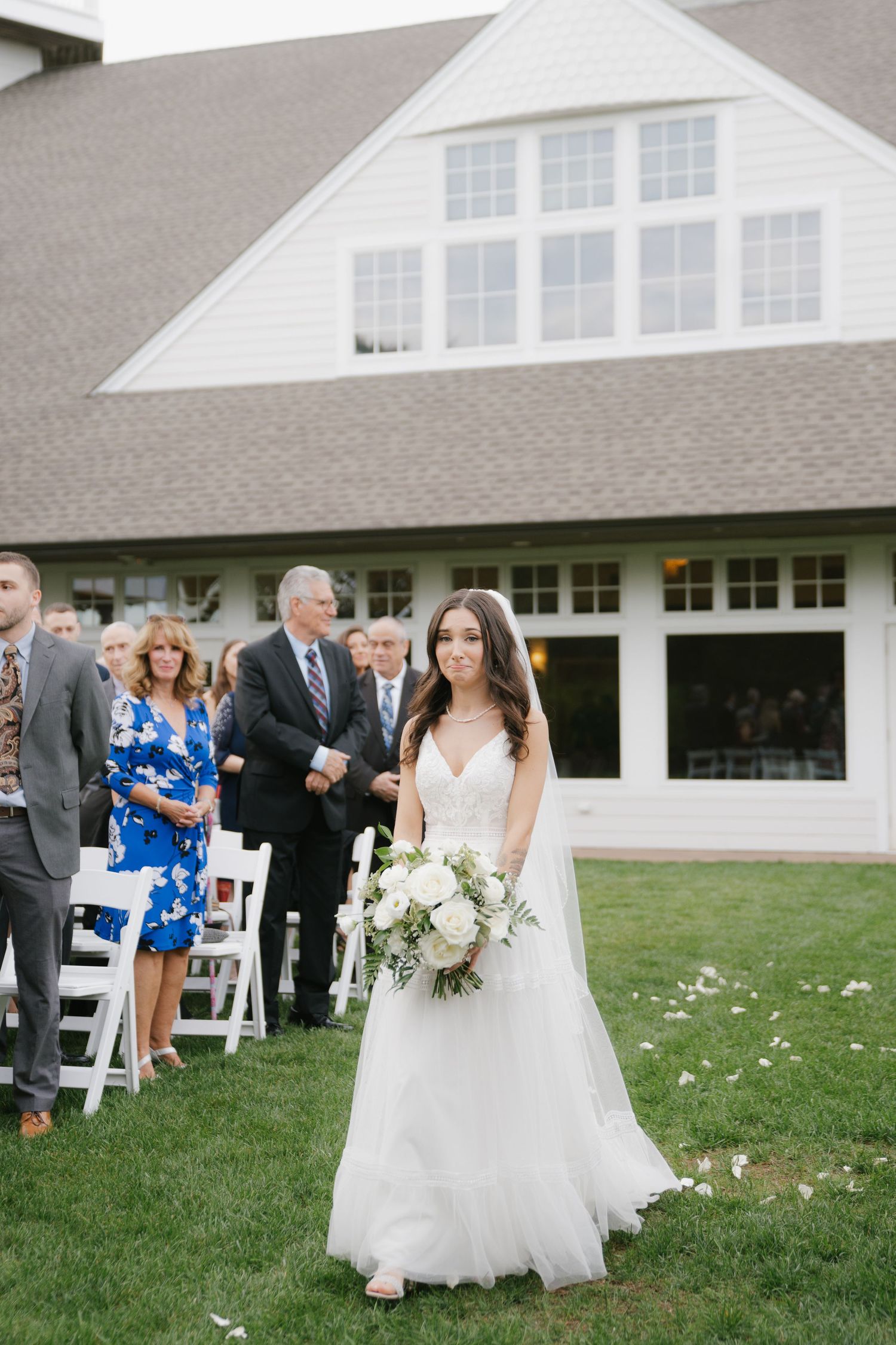 Wedding ceremony guests gather outside a white building with elegant windows on a grassy lawn.