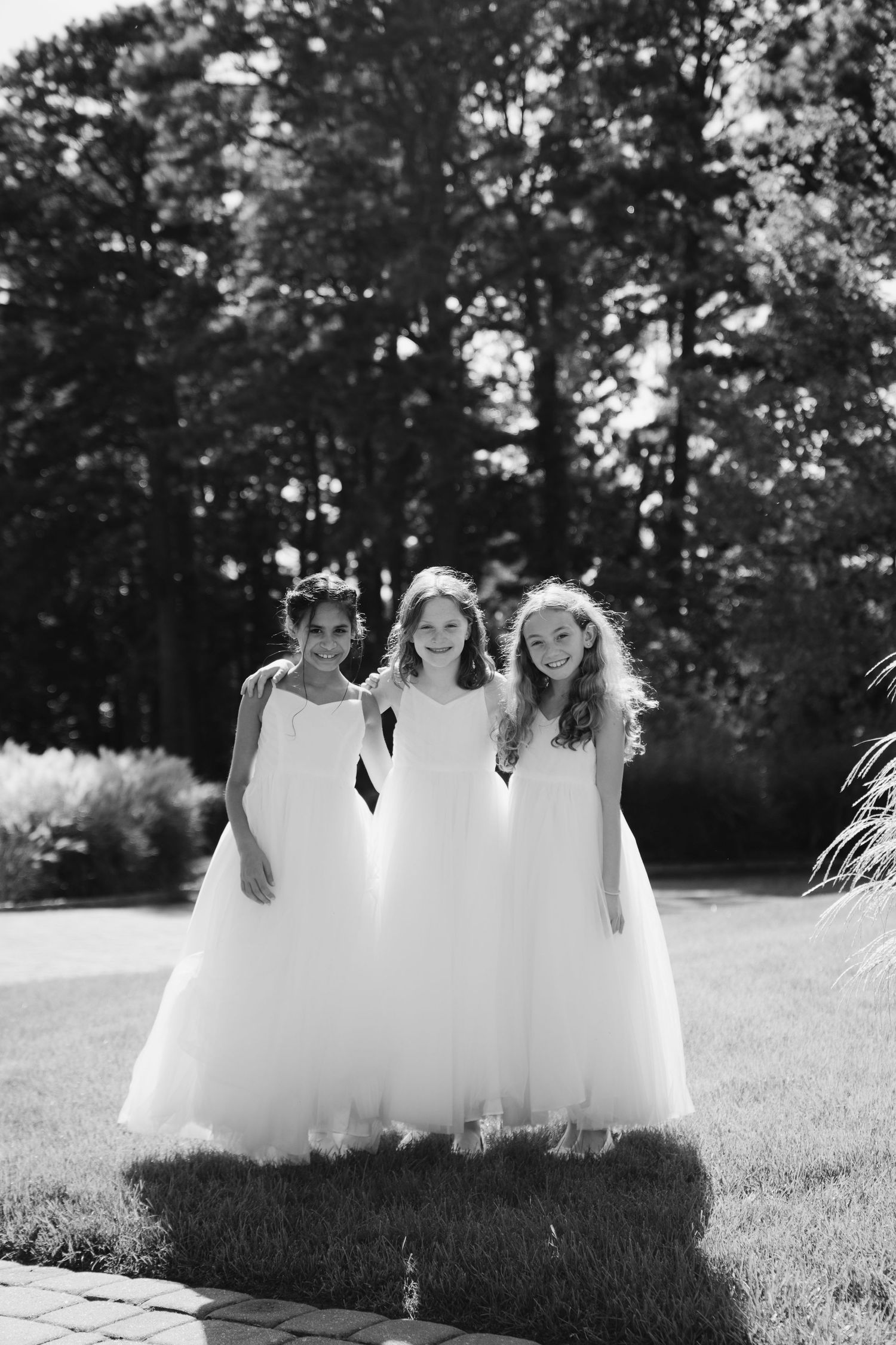Three young attendants in flowing white dresses stand together on a sunny lawn with trees in the background.