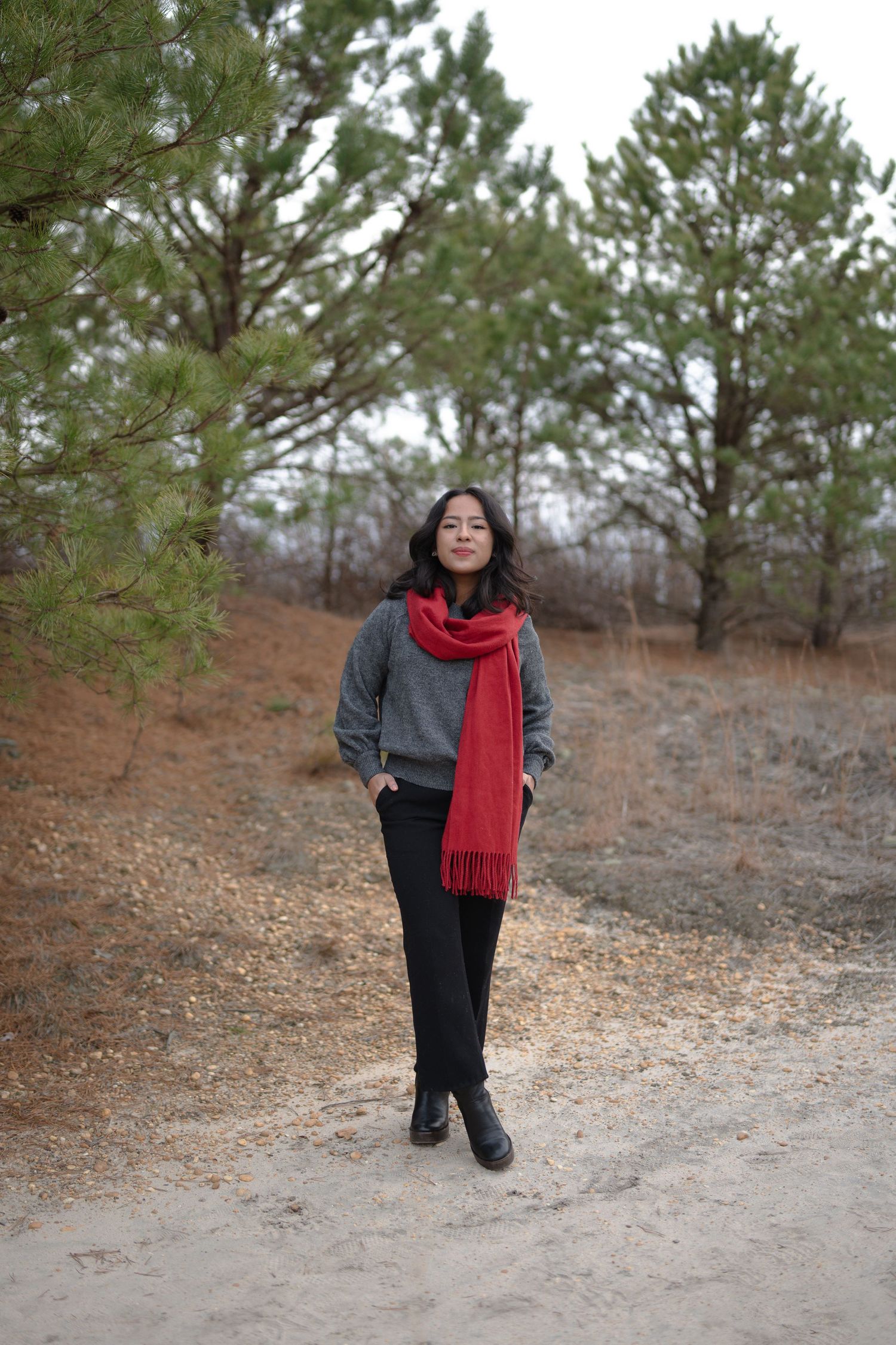 Person wearing a grey sweater and red scarf poses playfully outdoors in a winter landscape with pine trees.