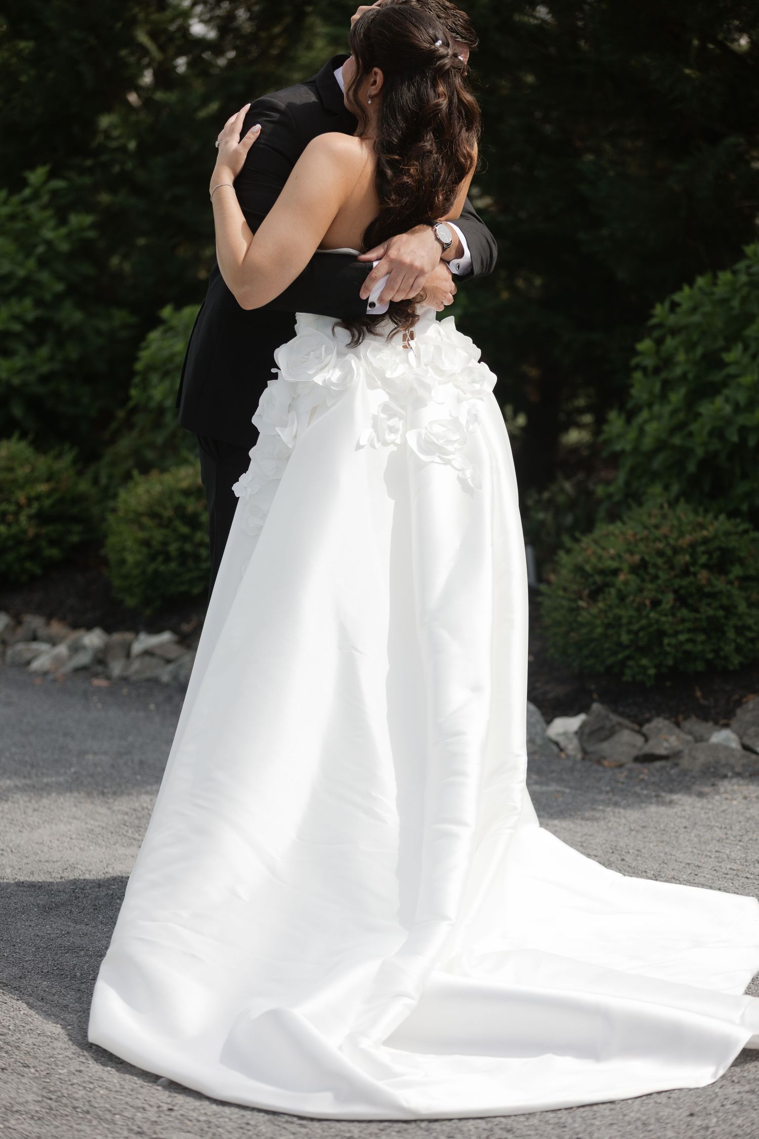 Bride and groom sharing romantic embrace outdoors, white wedding gown with floral details and long train.