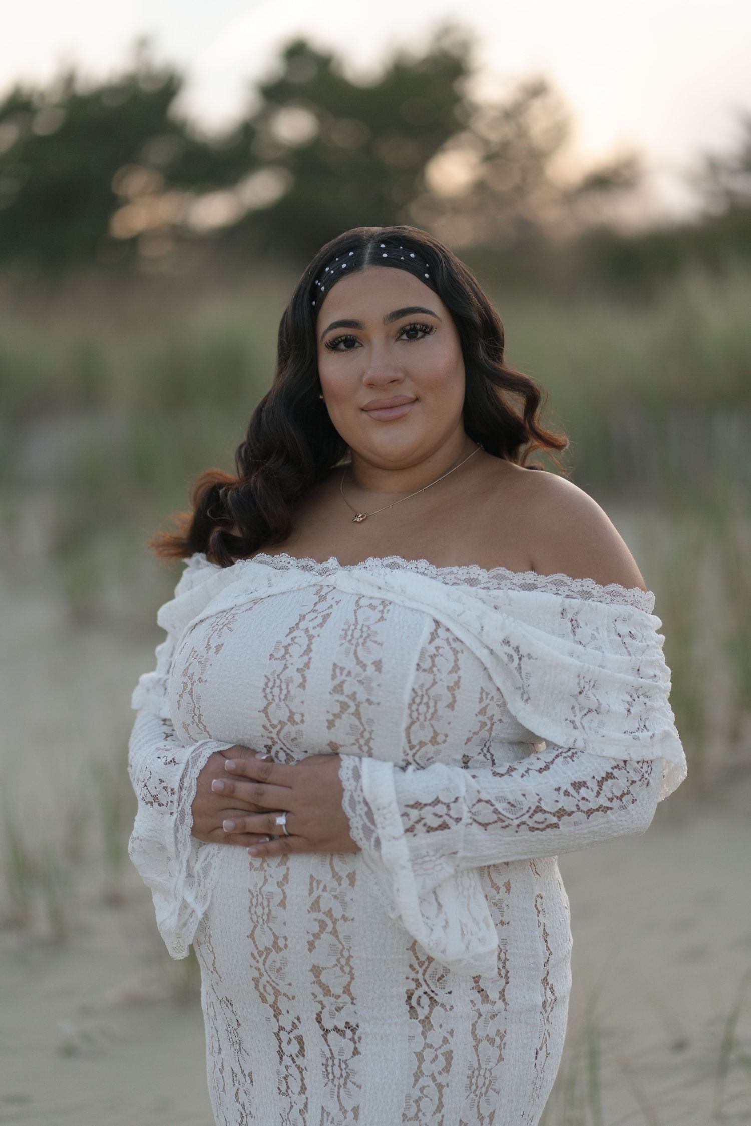 Woman in white off-shoulder lace dress poses on sandy beach at dusk, wearing a jeweled headband.