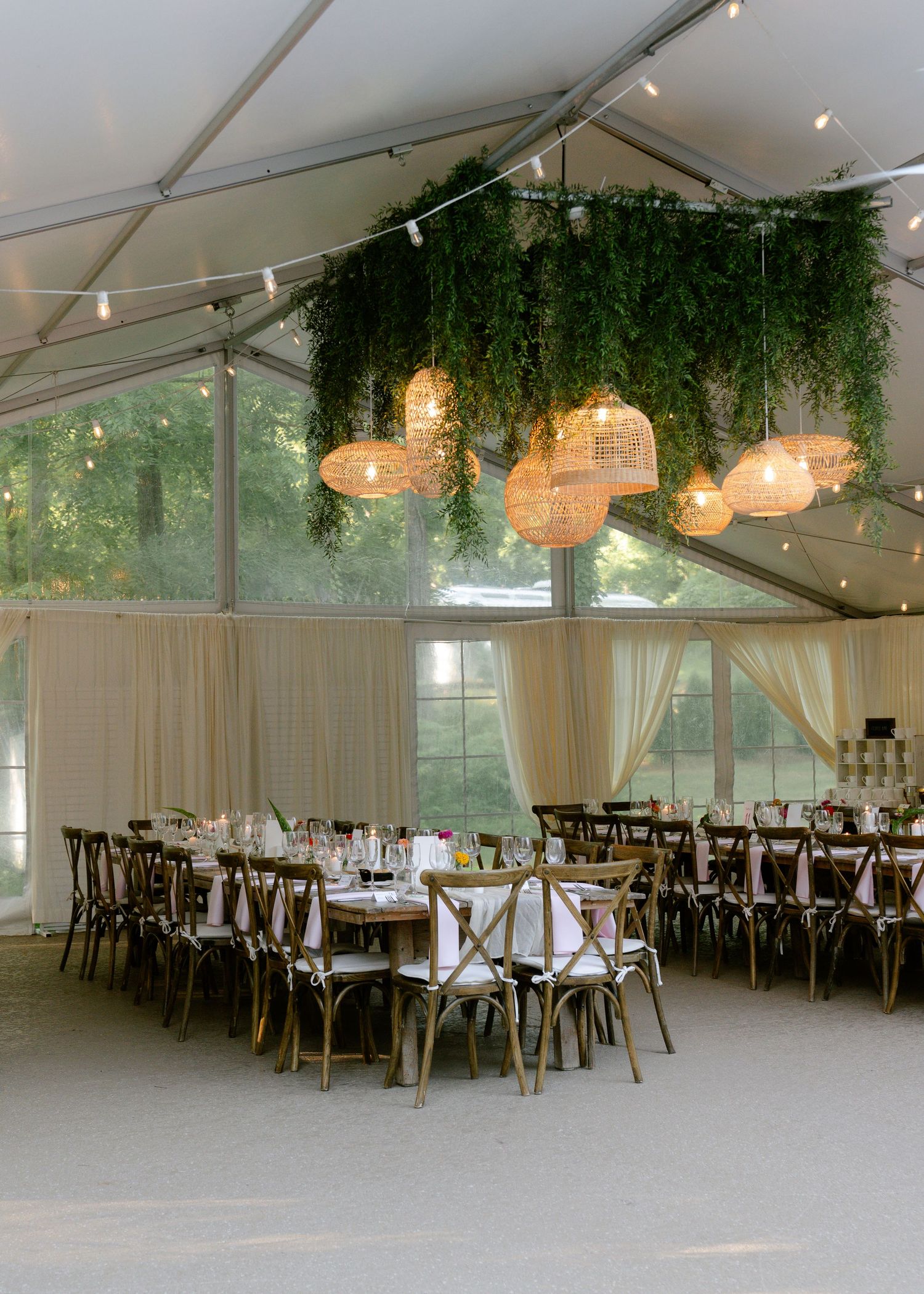 Elegant outdoor wedding tent reception with rattan pendant lights, lush greenery ceiling installation, round tables, and cross-back chairs.