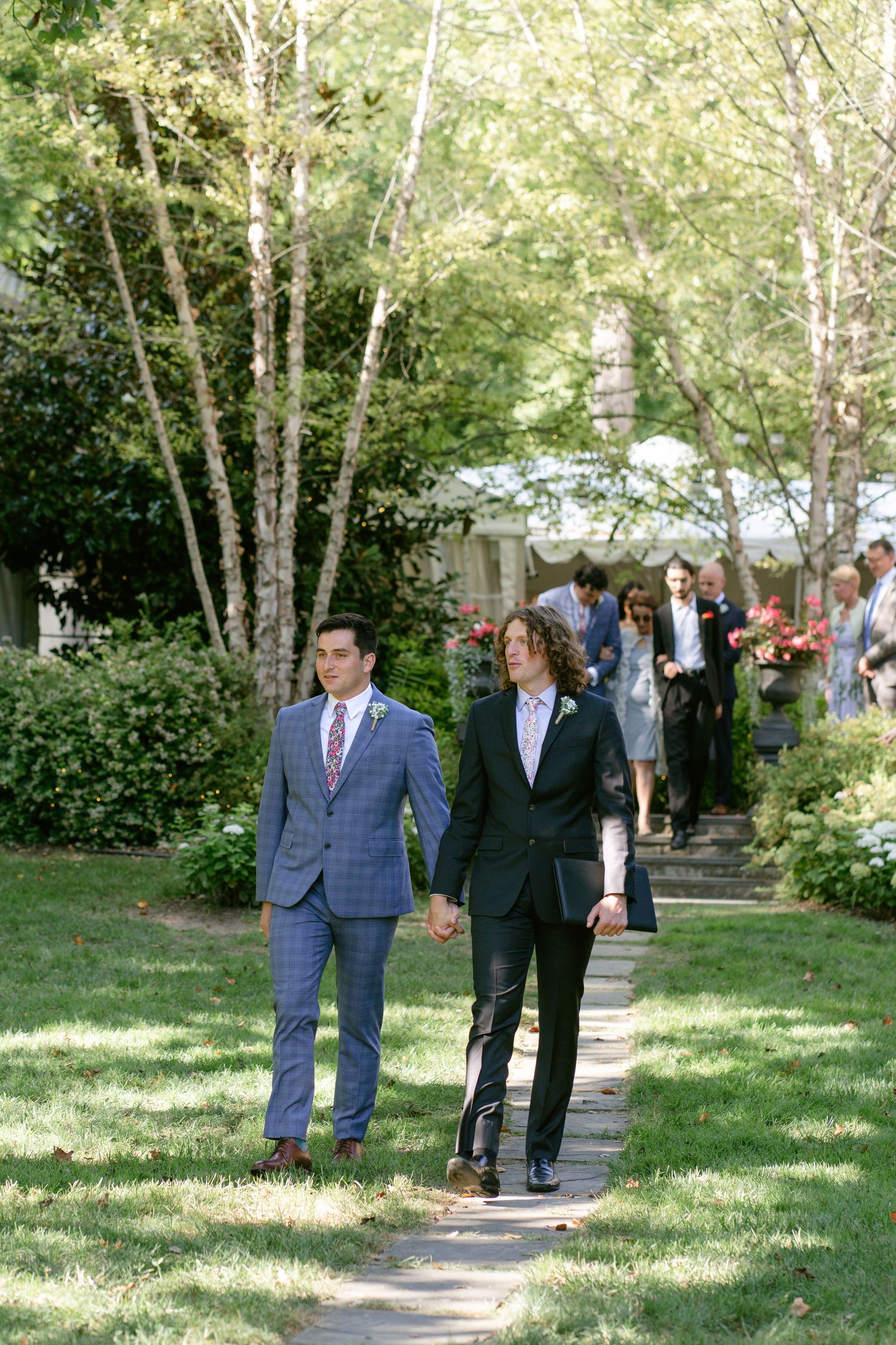 Wedding party members walking down tree-lined pathway in formal attire.