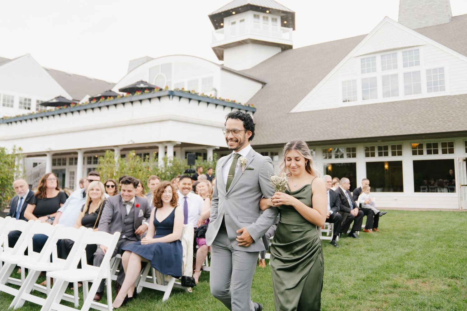 A wedding party walks down the aisle on a lawn with white chairs and a beautiful white building in the background.