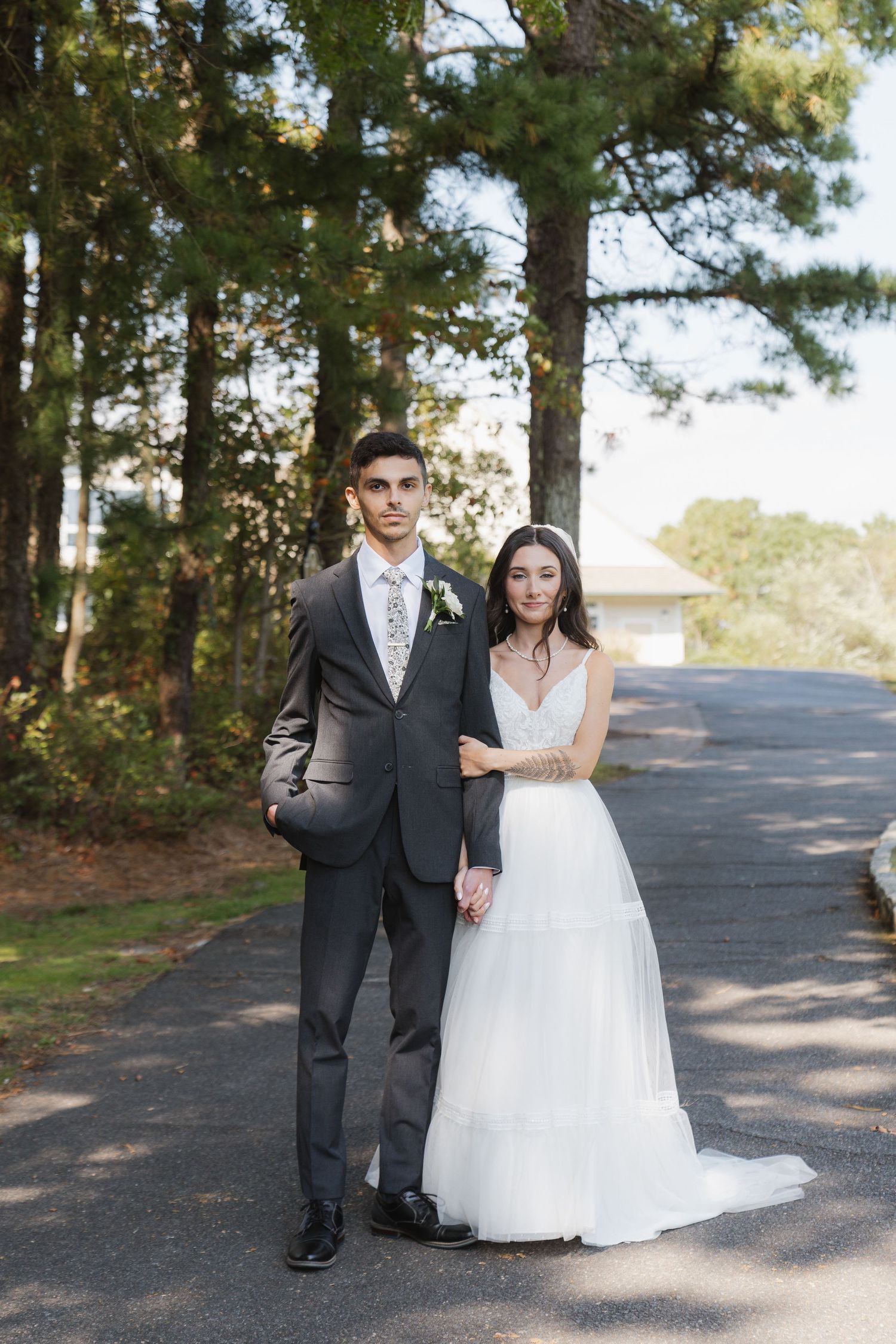 A couple in wedding attire stands together on a tree-lined path during their outdoor wedding photoshoot.