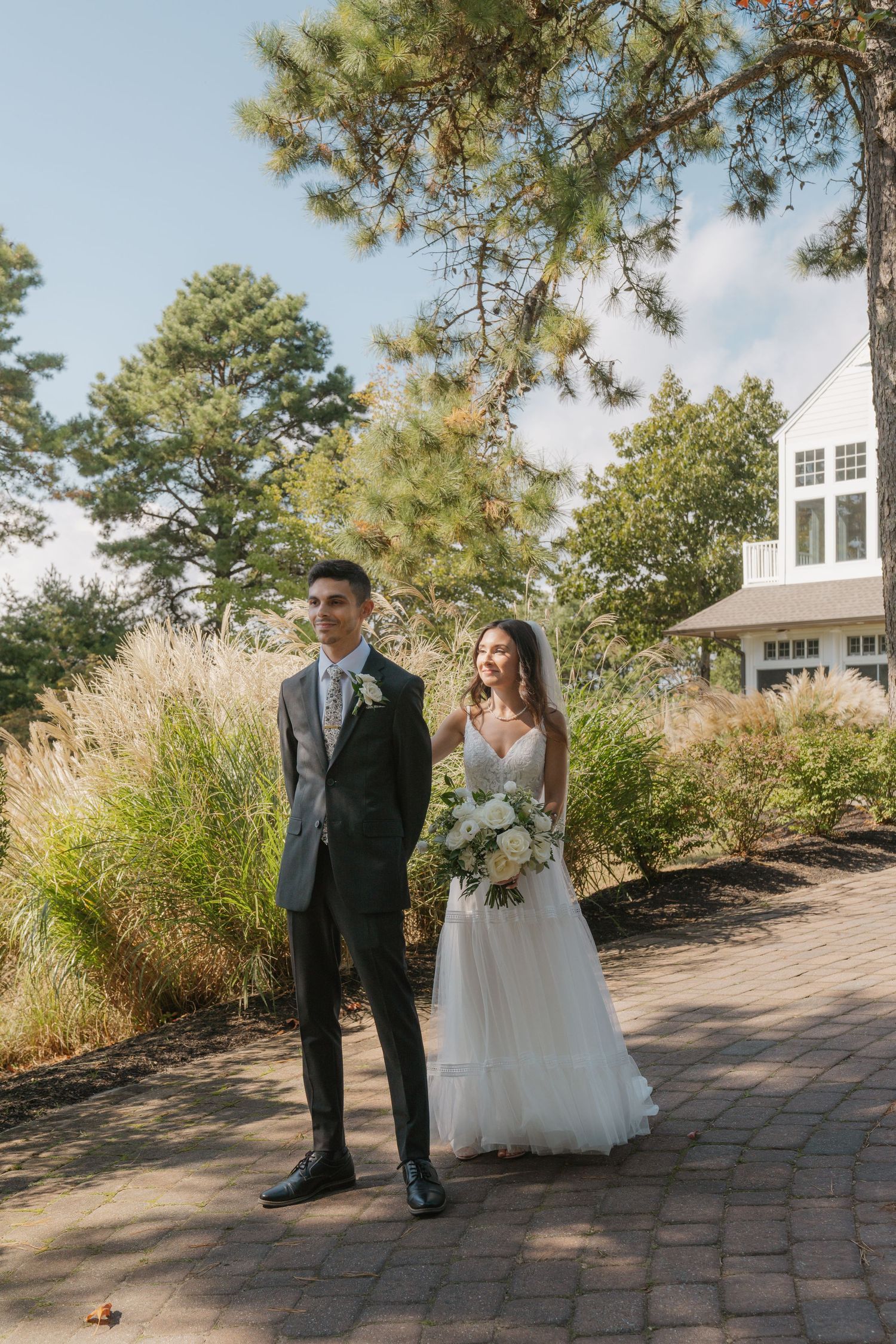 Wedding couple stands on brick patio surrounded by tall ornamental grasses and pine trees on sunny day.