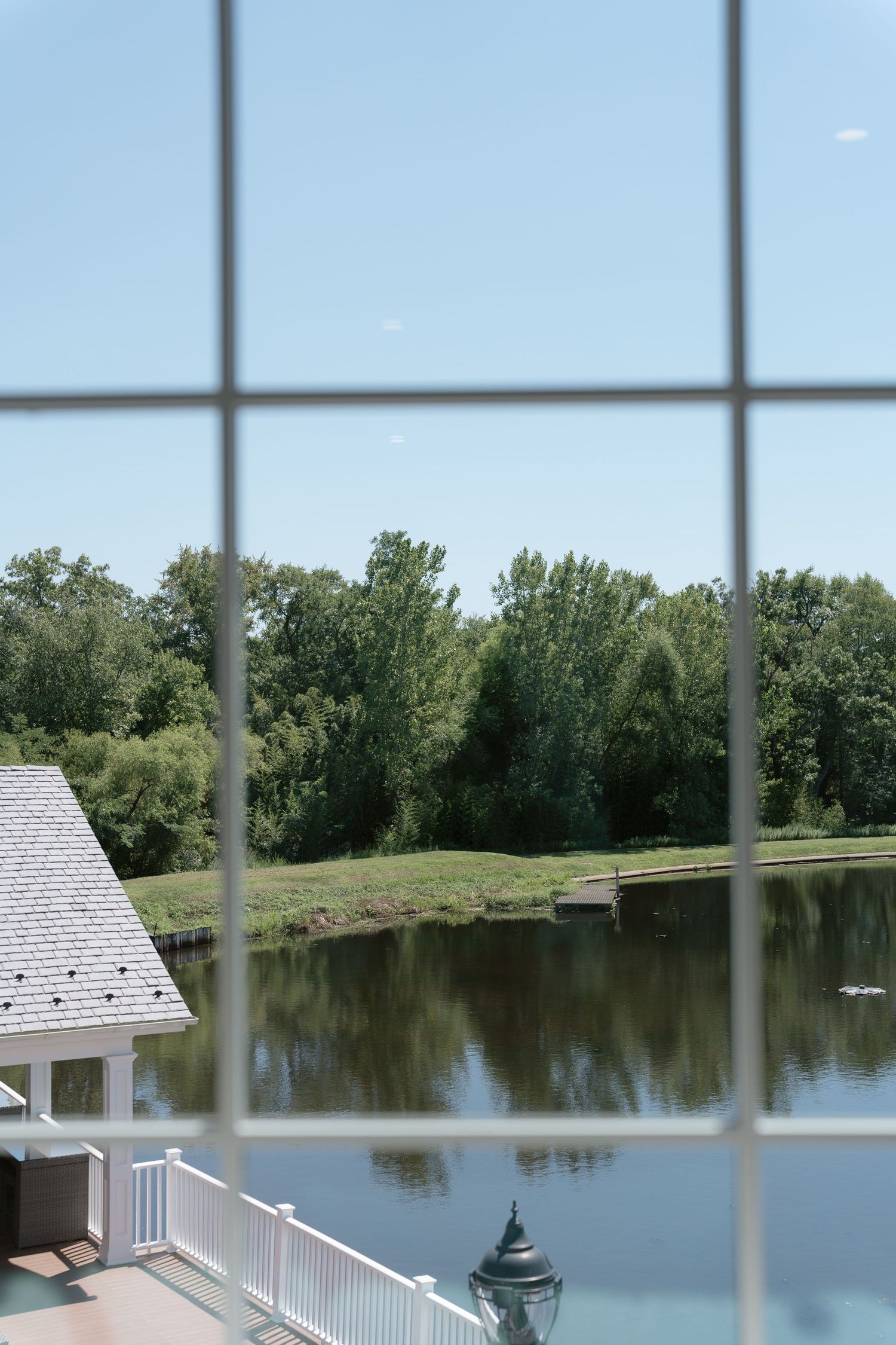 View through large windows of a serene pond surrounded by lush green trees and a white railed deck on a sunny day.