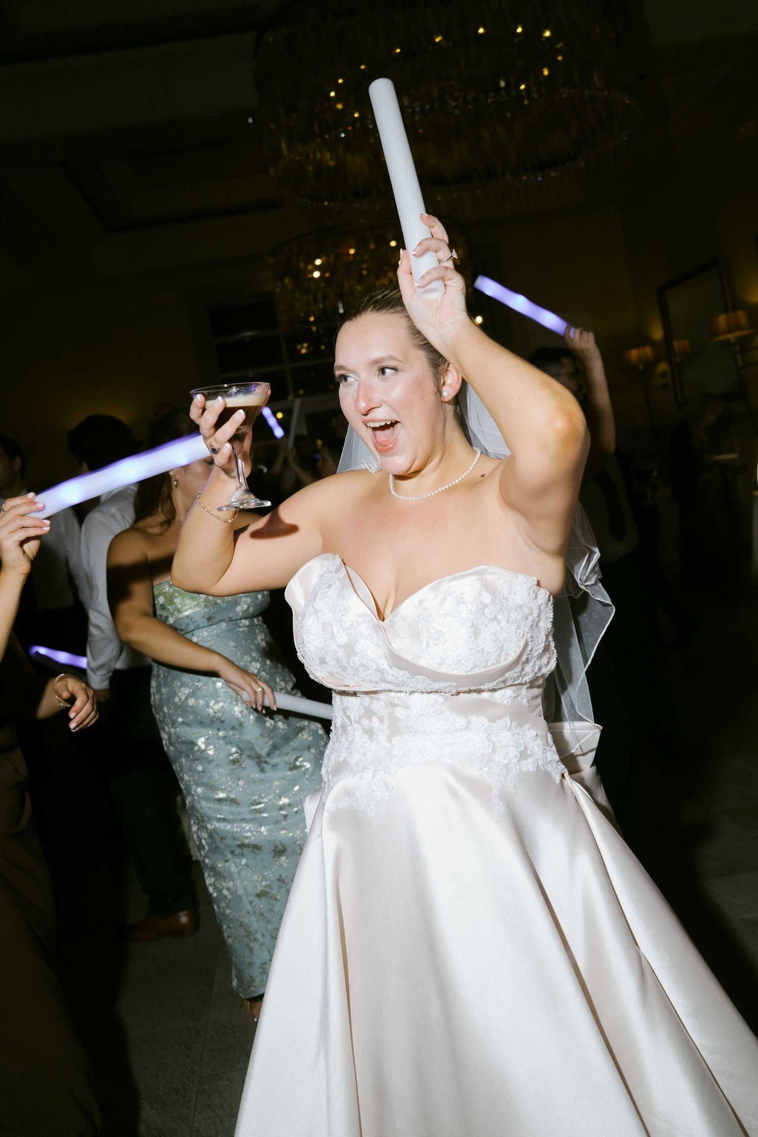 Excited bride dances with glow stick at indoor wedding reception celebration.