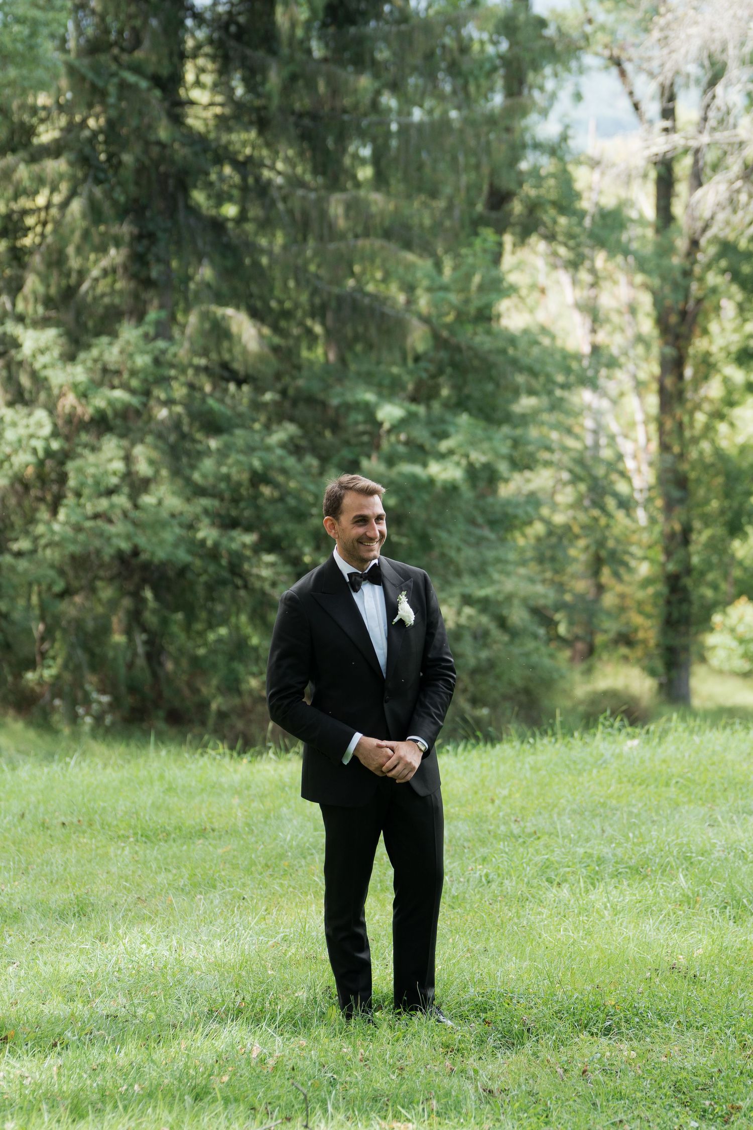 Groom in black tuxedo with bow tie and boutonniere smiling outdoors on green grass with trees behind.