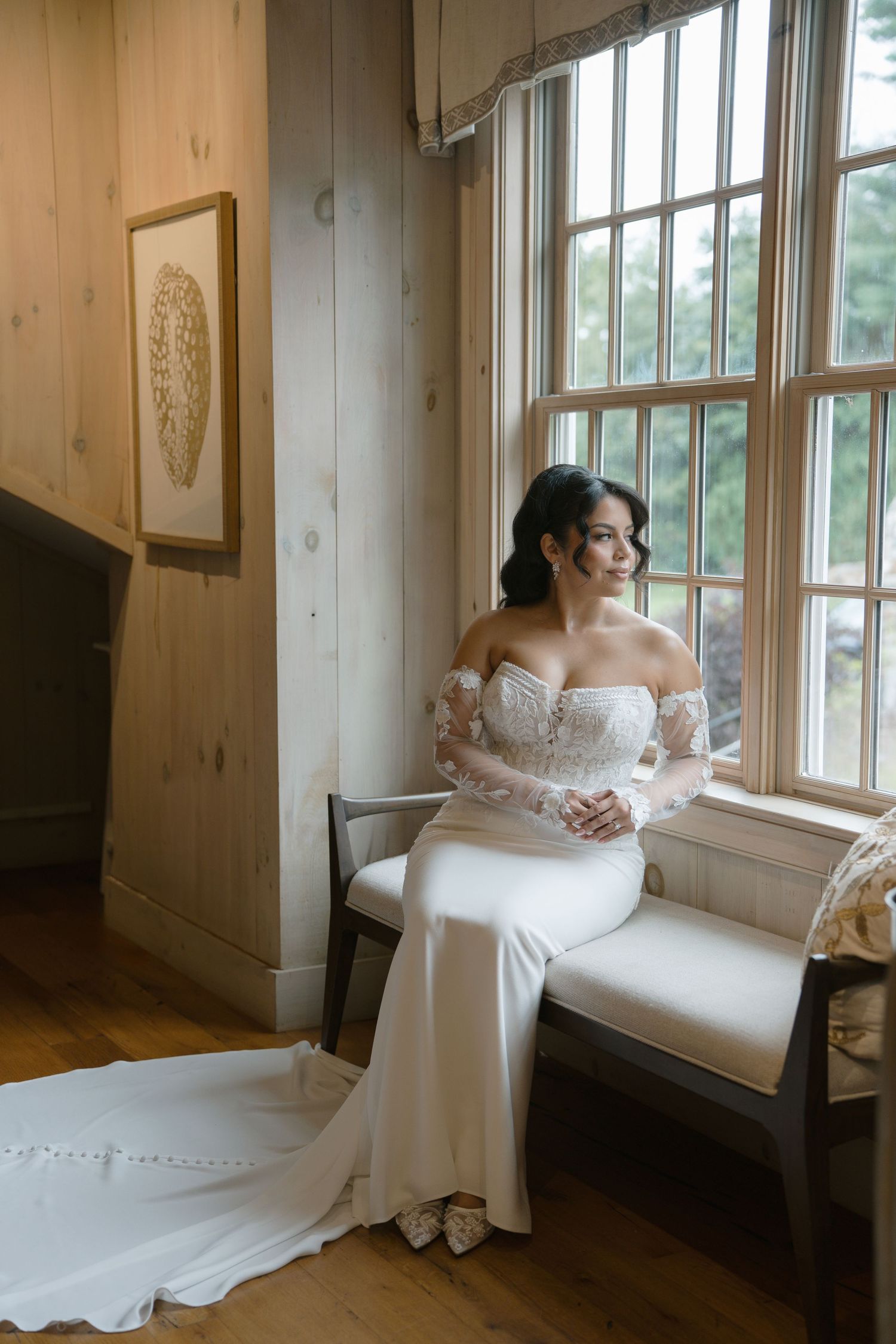 Bride in white off-shoulder gown with lace sleeves sits on bench by large window in rustic wood-paneled room.