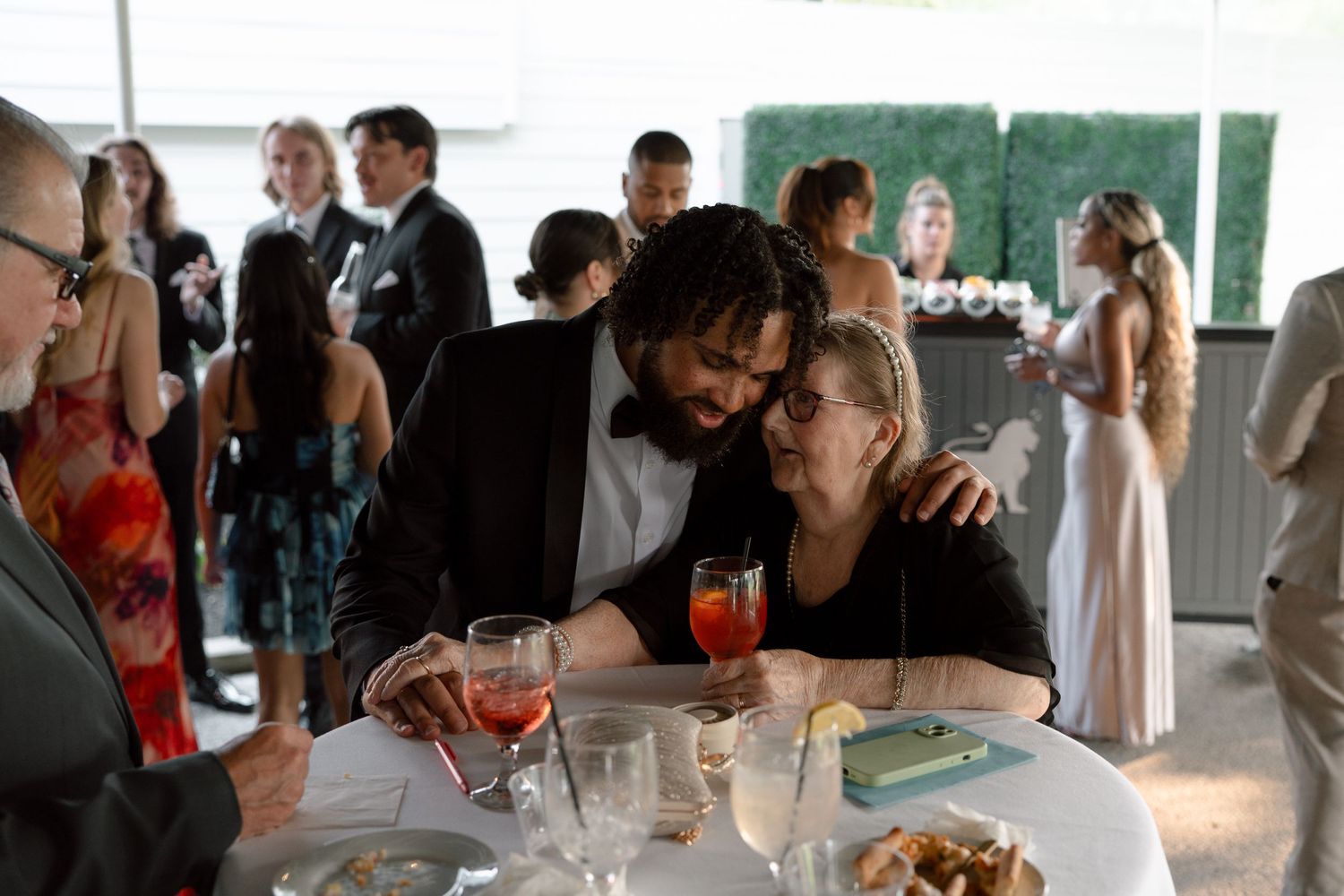 Couple sharing an intimate moment at a wedding reception, sitting at a table with cocktails and guests mingling in background.
