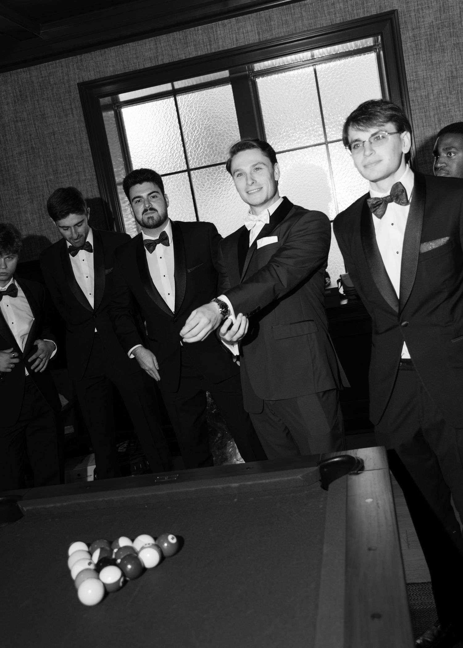 Men in tuxedos with bow ties gather around a pool table, smiling and posing for a black and white photo.