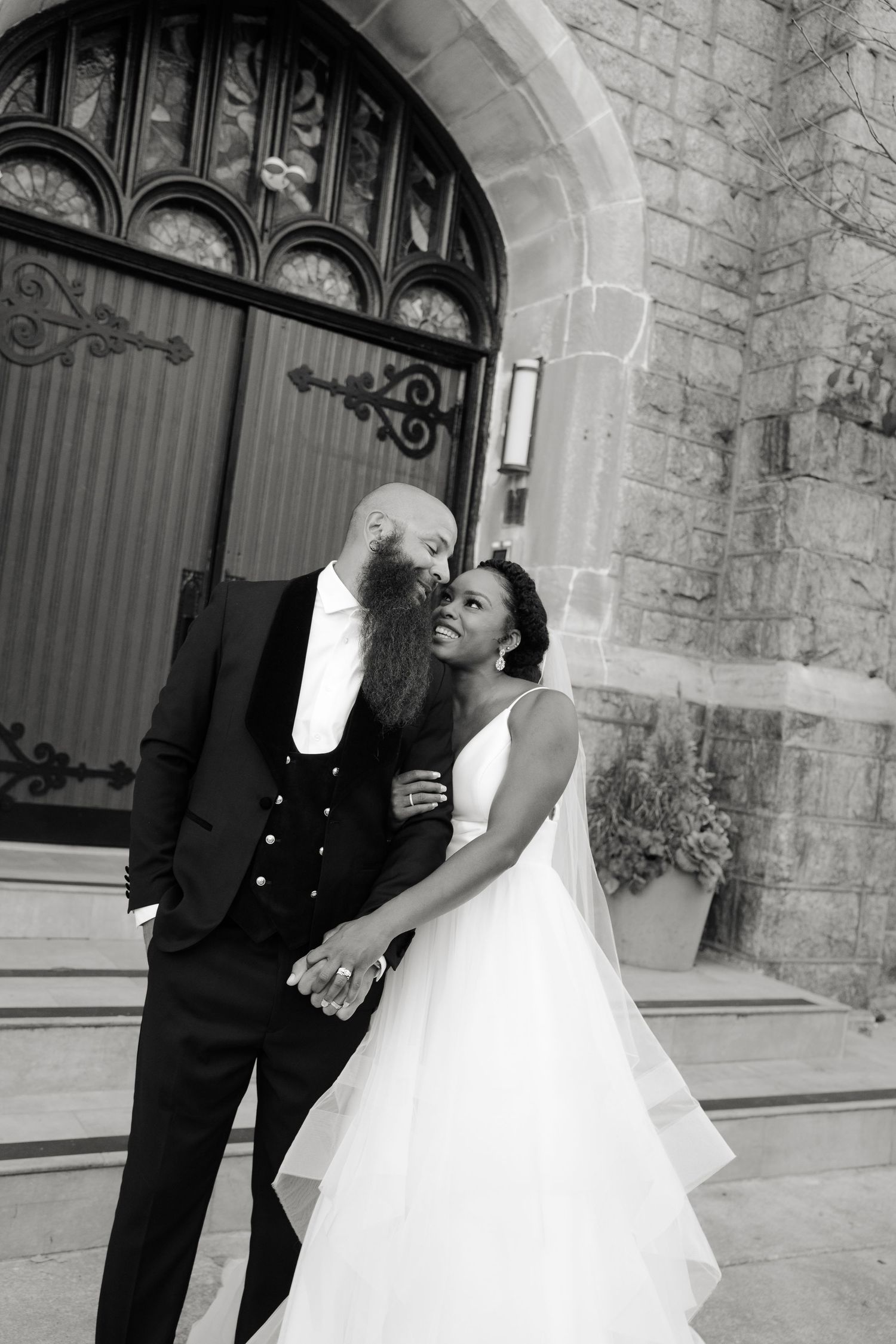 Black and white wedding photo of a bearded groom and bride in white gown posing before ornate church doors.
