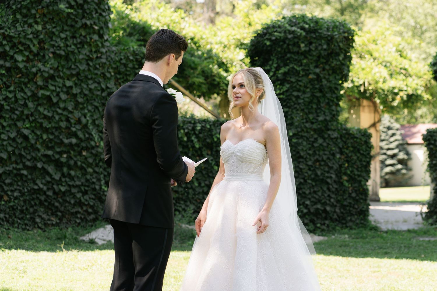 Groom reads vows to blonde bride in white strapless wedding gown and veil outdoors among lush green hedges.