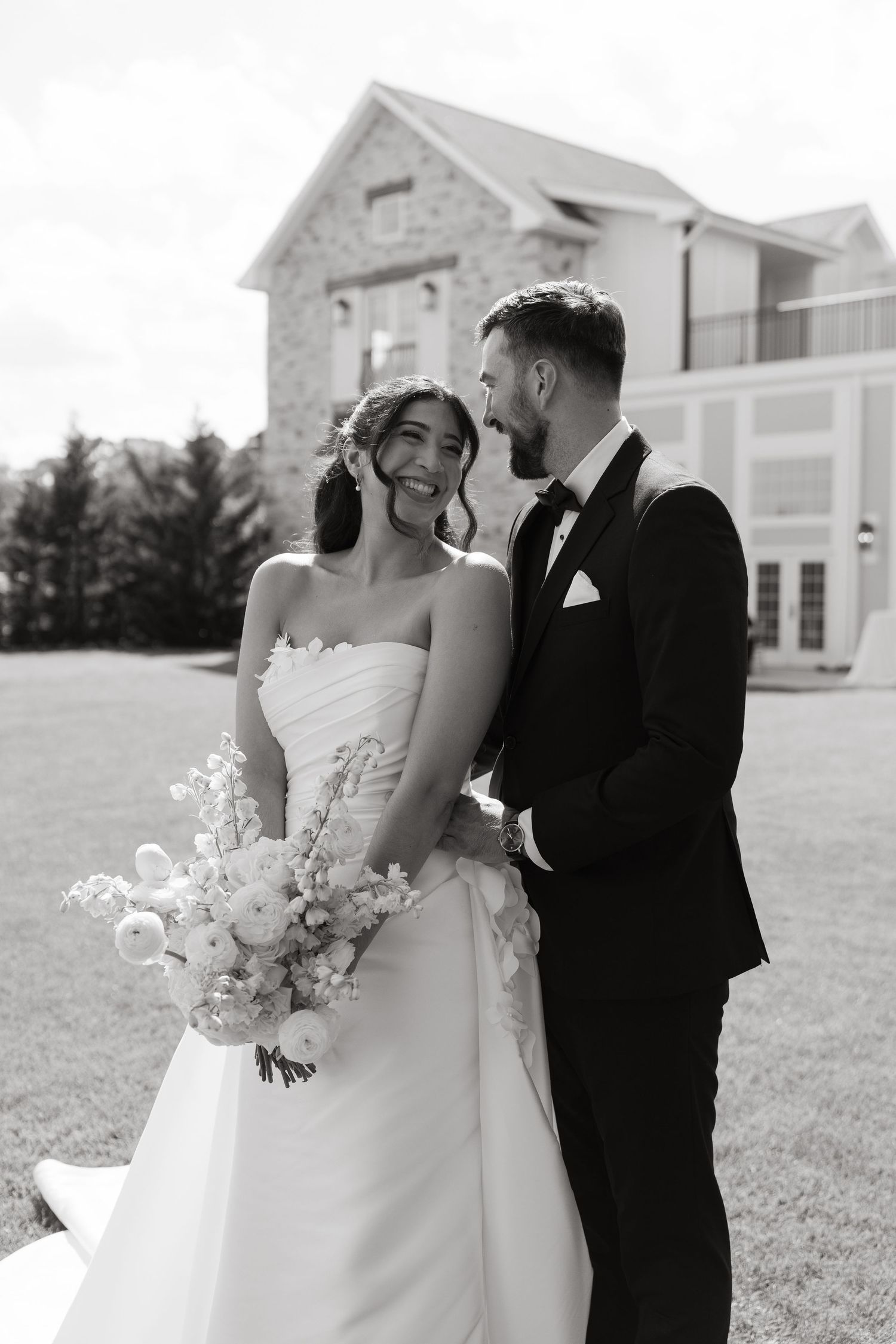 Black and white photo of smiling bride holding bouquet with groom in tuxedo standing outside stone estate.