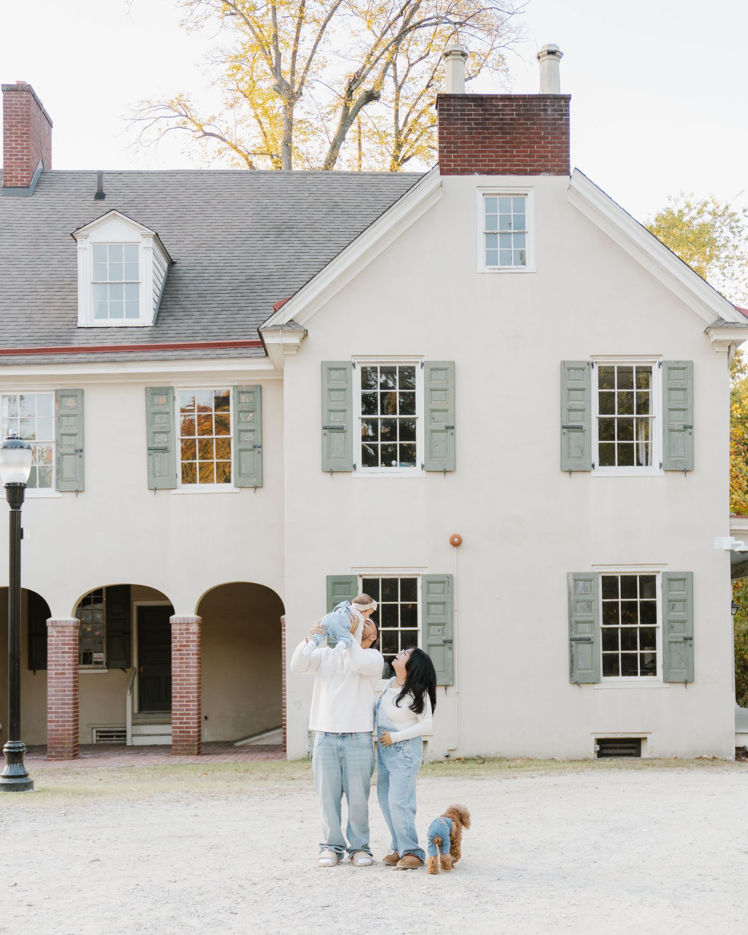 A couple and their dog stand in front of a white colonial style house with gray shutters and brick archways.