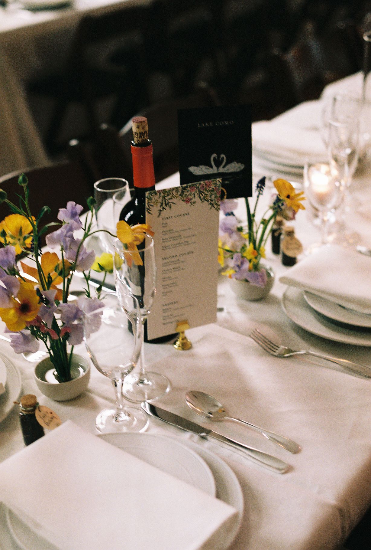 Elegant wedding table setting with white linens, purple and yellow flowers in small vases, wine glasses, and menu cards.