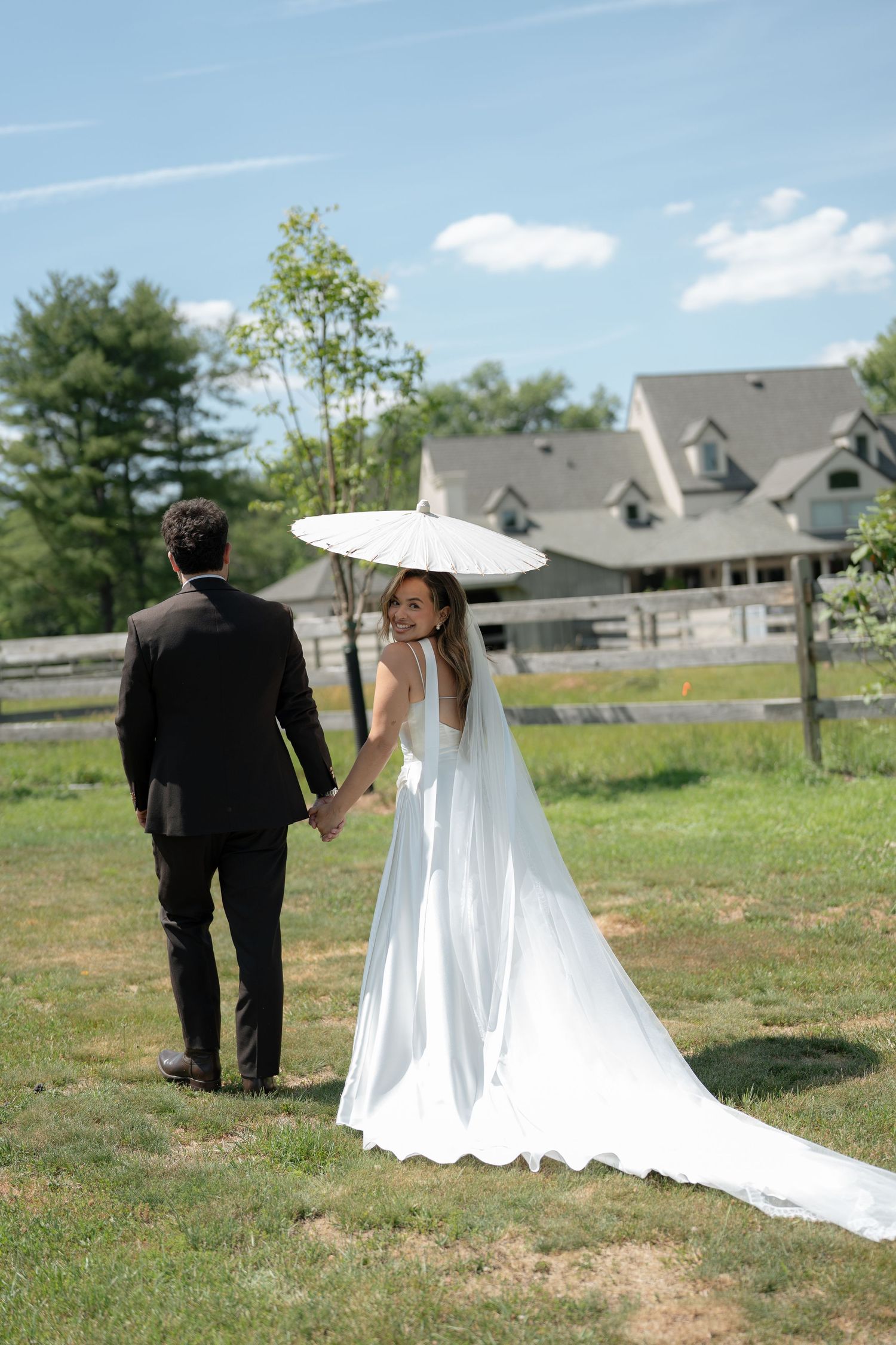Bride in white gown holding a parasol and groom in black suit walk hand-in-hand across green lawn.