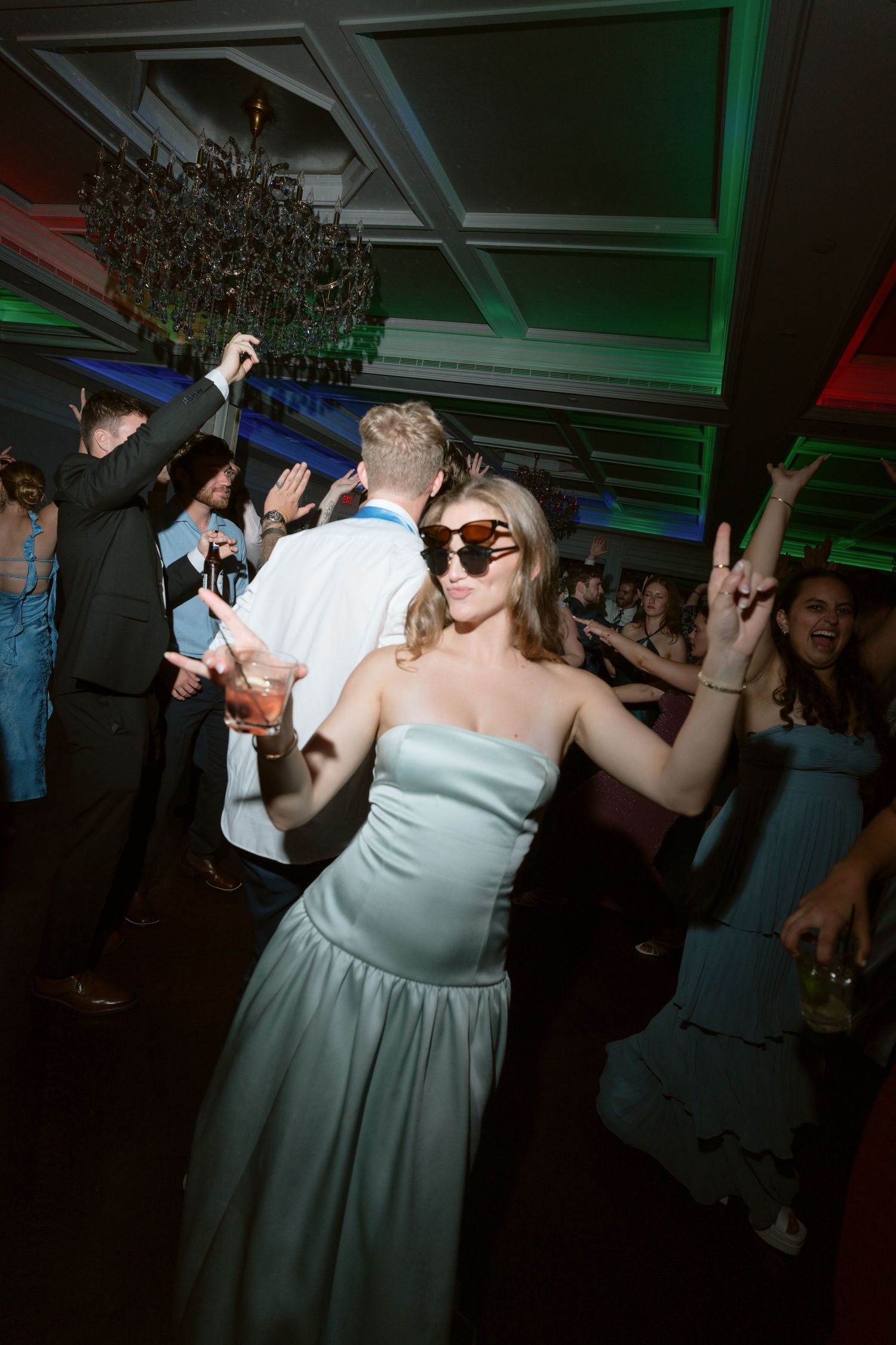 Bridesmaid in blue gown wearing sunglasses dancing at lively wedding reception with colorful lights.