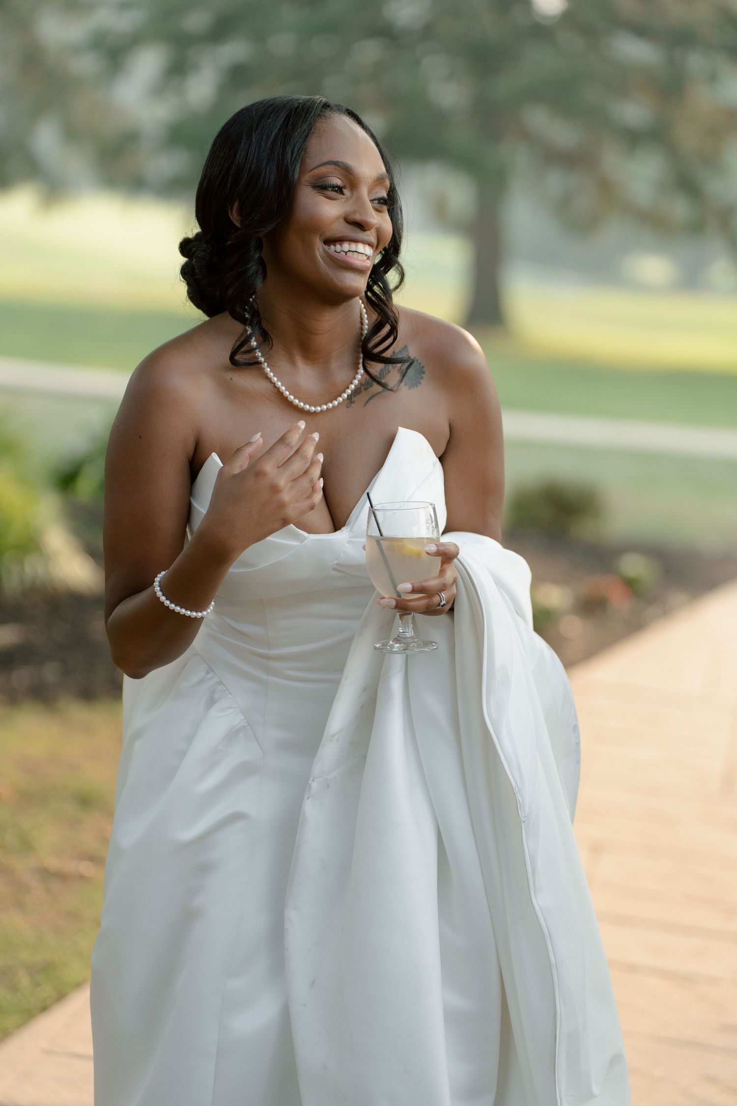 Smiling Black bride in strapless white gown holding champagne glass outdoors, wearing pearl necklace and bracelet.