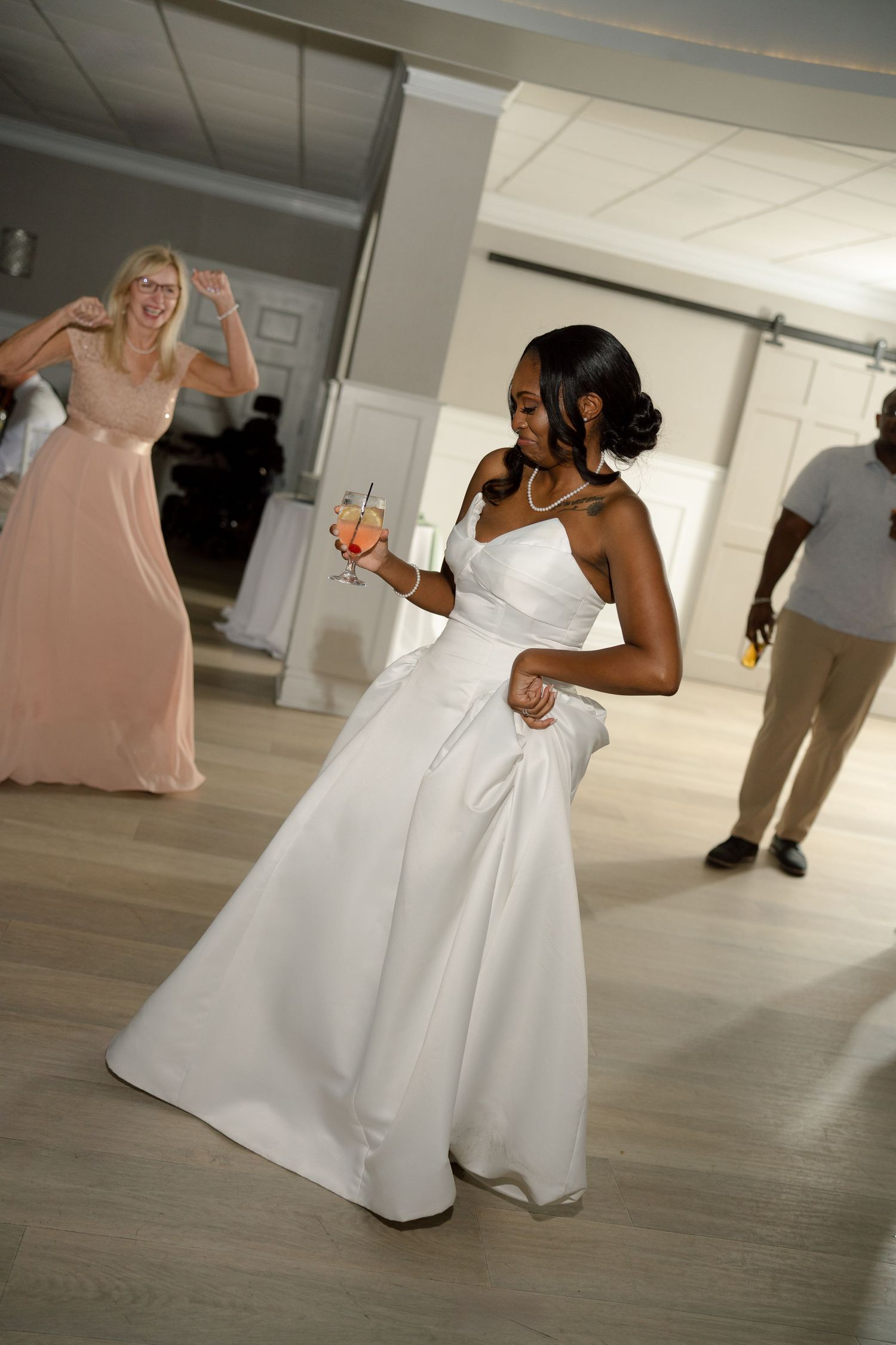 Radiant bride in white gown dances joyfully holding cocktail at wedding reception celebration.