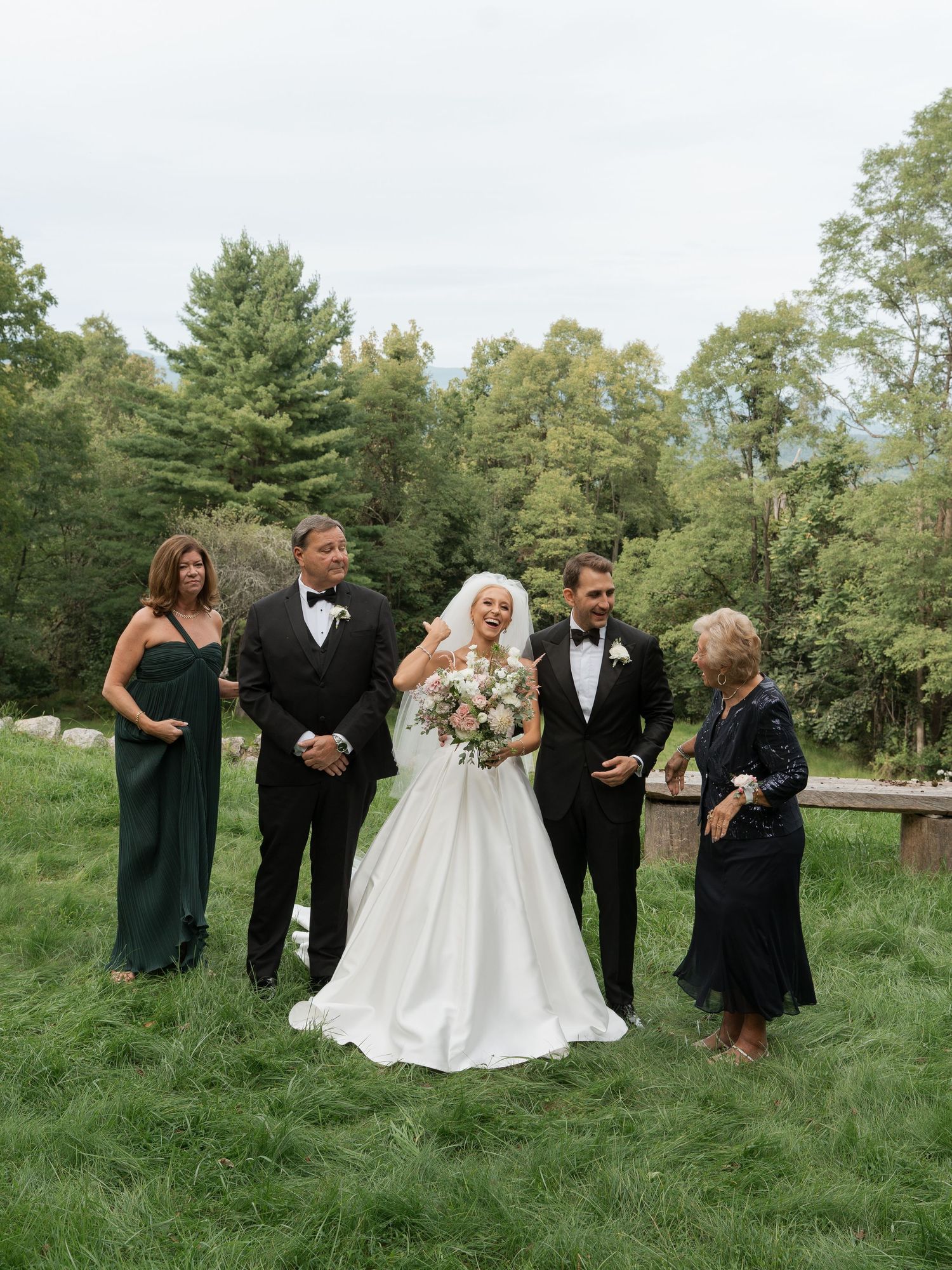 Bride in white gown with groom and family members in formal attire posing outdoors on green lawn with forest backdrop.
