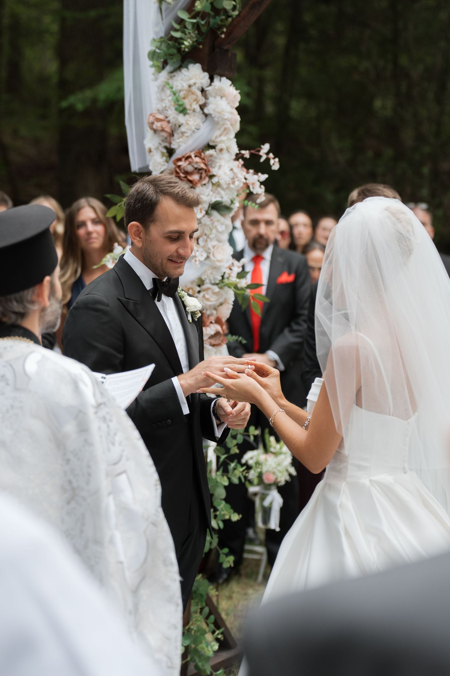 Groom smiles while placing wedding ring on brides hand at floral outdoor ceremony altar.