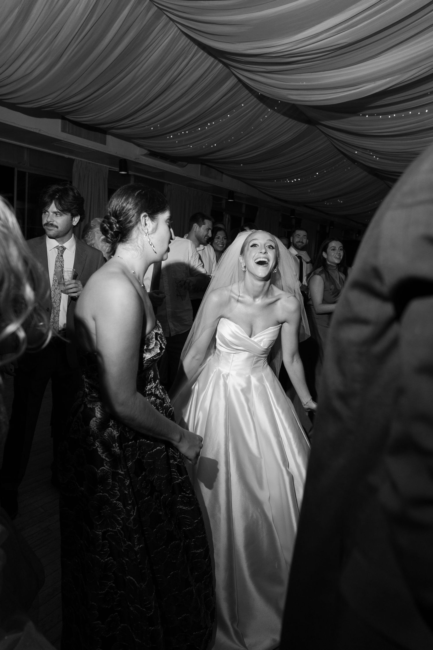 Black and white photo of bride in satin gown dancing joyfully among guests at wedding reception.
