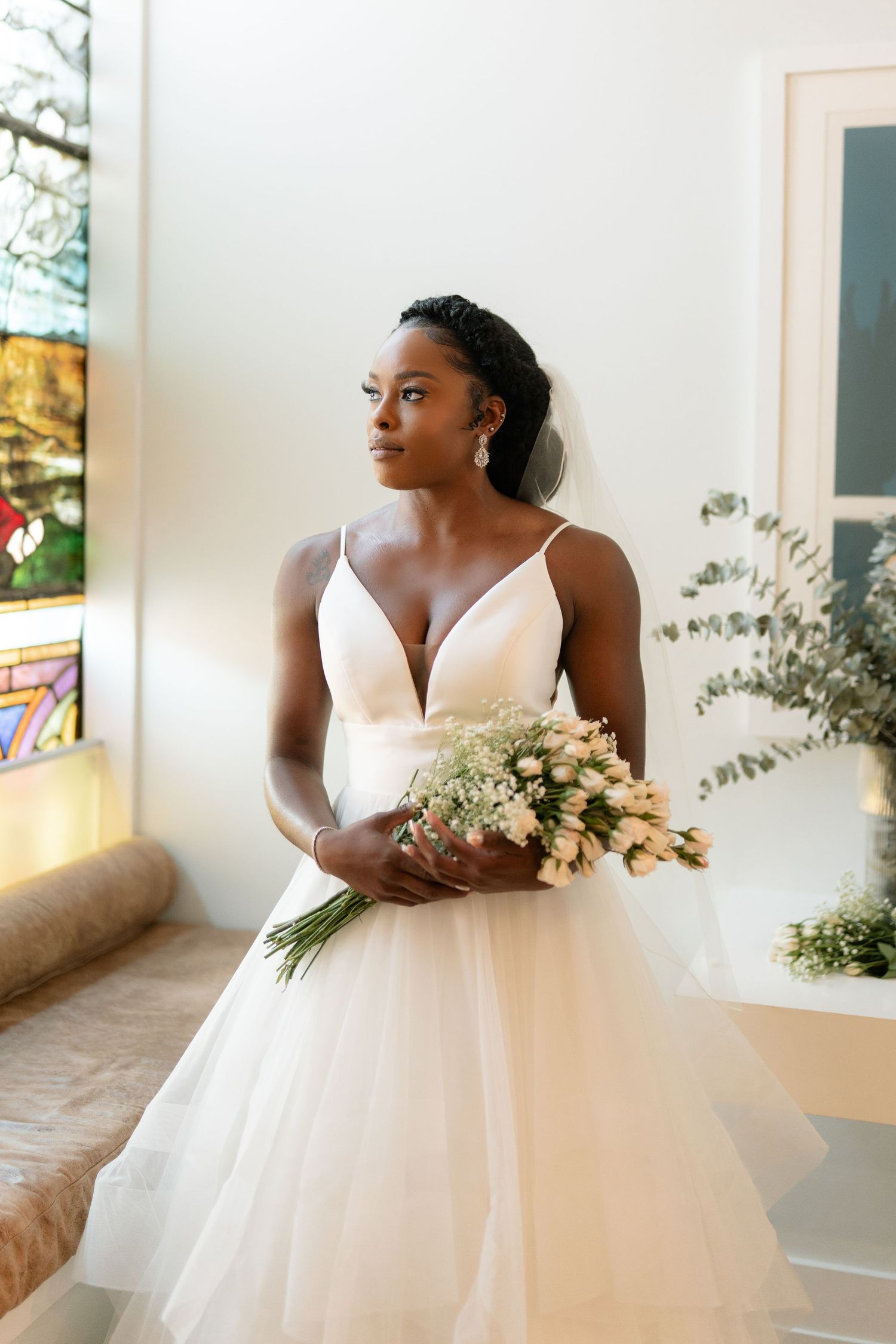Elegant bride in white plunging neckline ballgown holds delicate bouquet of white flowers near window.