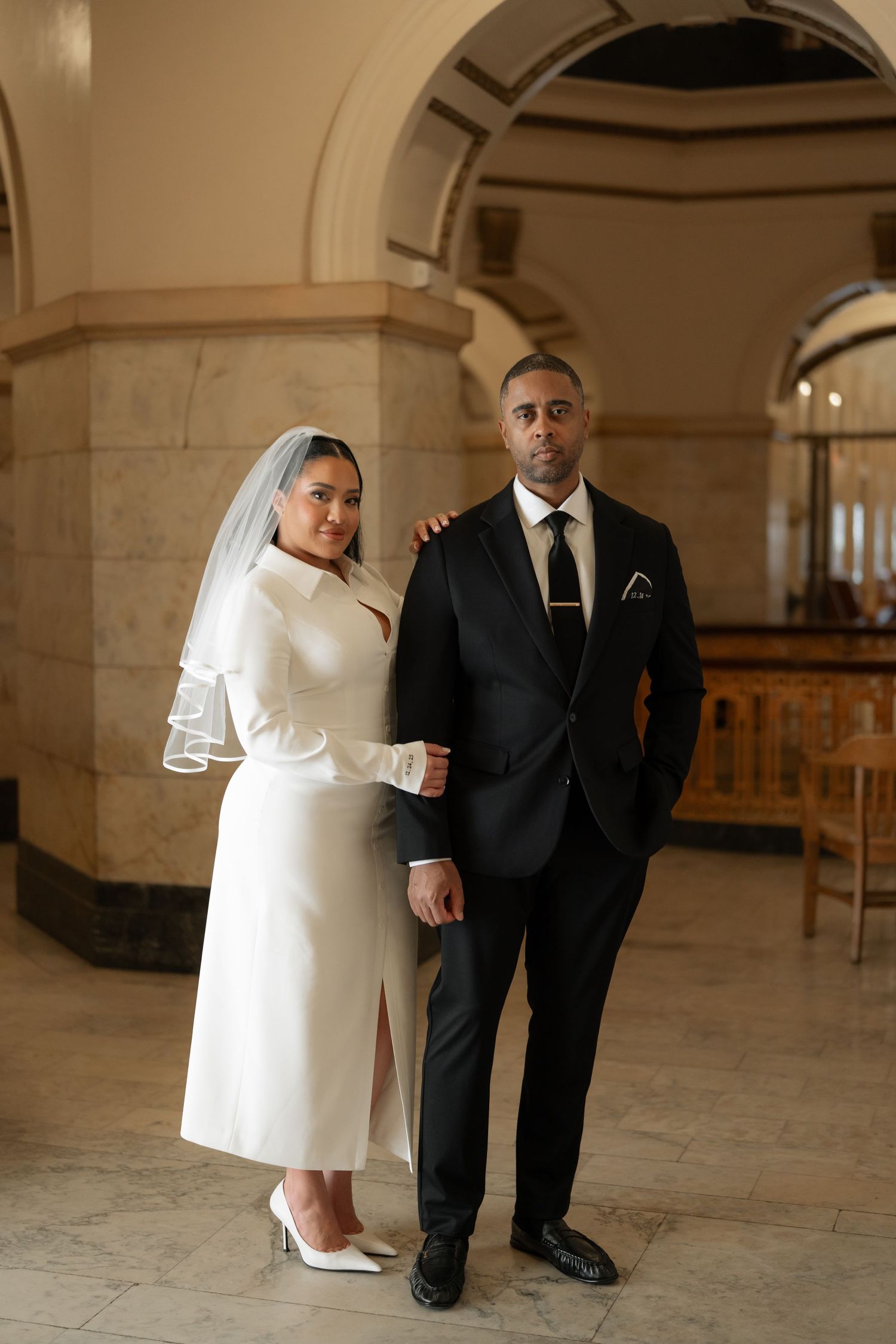 A bride in white dress and groom in black suit pose together under stone archways.