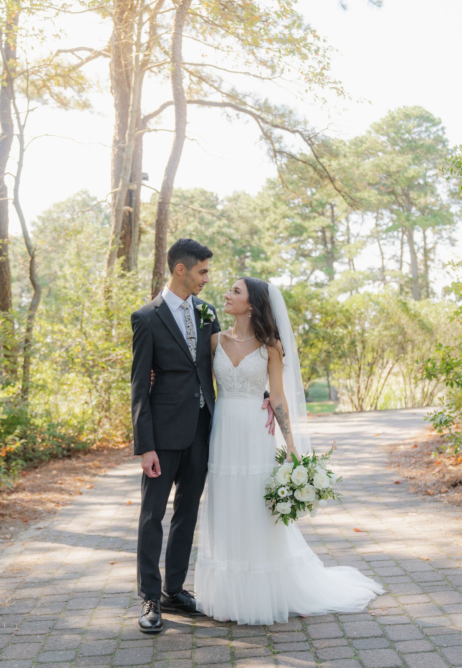 A newlywed couple stands together on a stone path surrounded by tall pine trees on their wedding day.