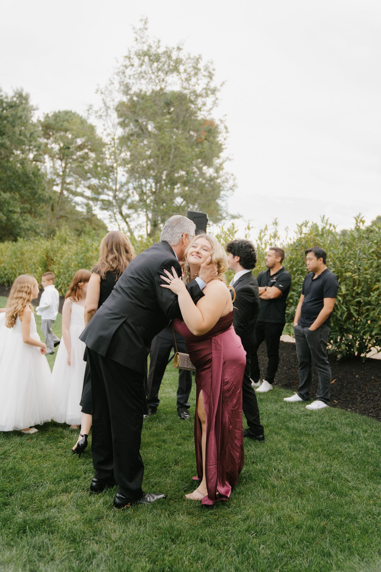 A couple shares a romantic moment on a grassy lawn while wedding guests in formal attire look on during an outdoor celebration.