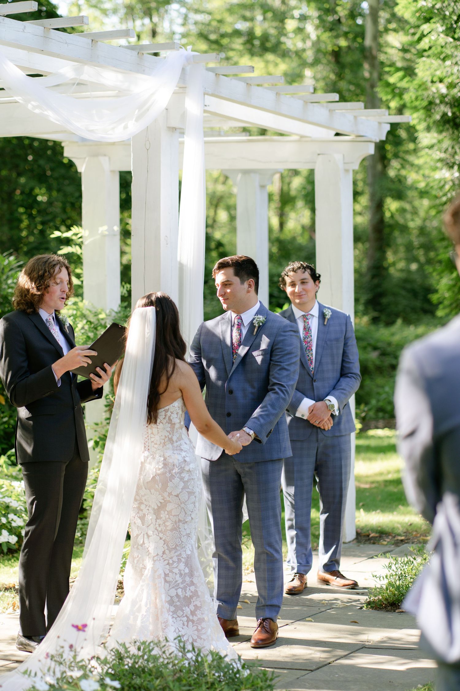 Wedding celebration under a white pergola with guests and wedding party in blue suits.