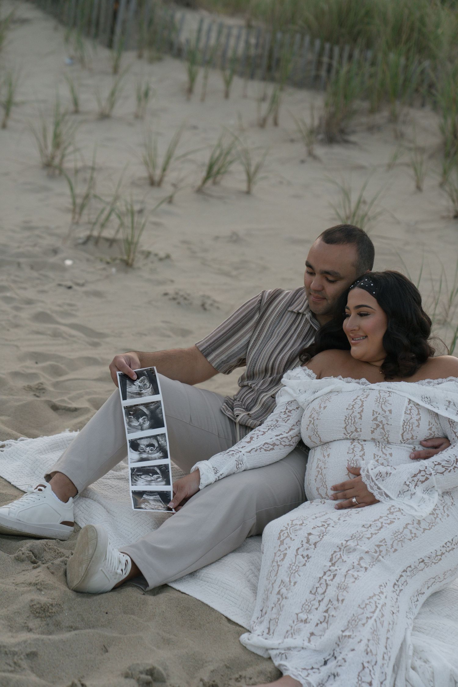 Expecting couple sitting on sandy beach dunes looking at ultrasound photos together tenderly.